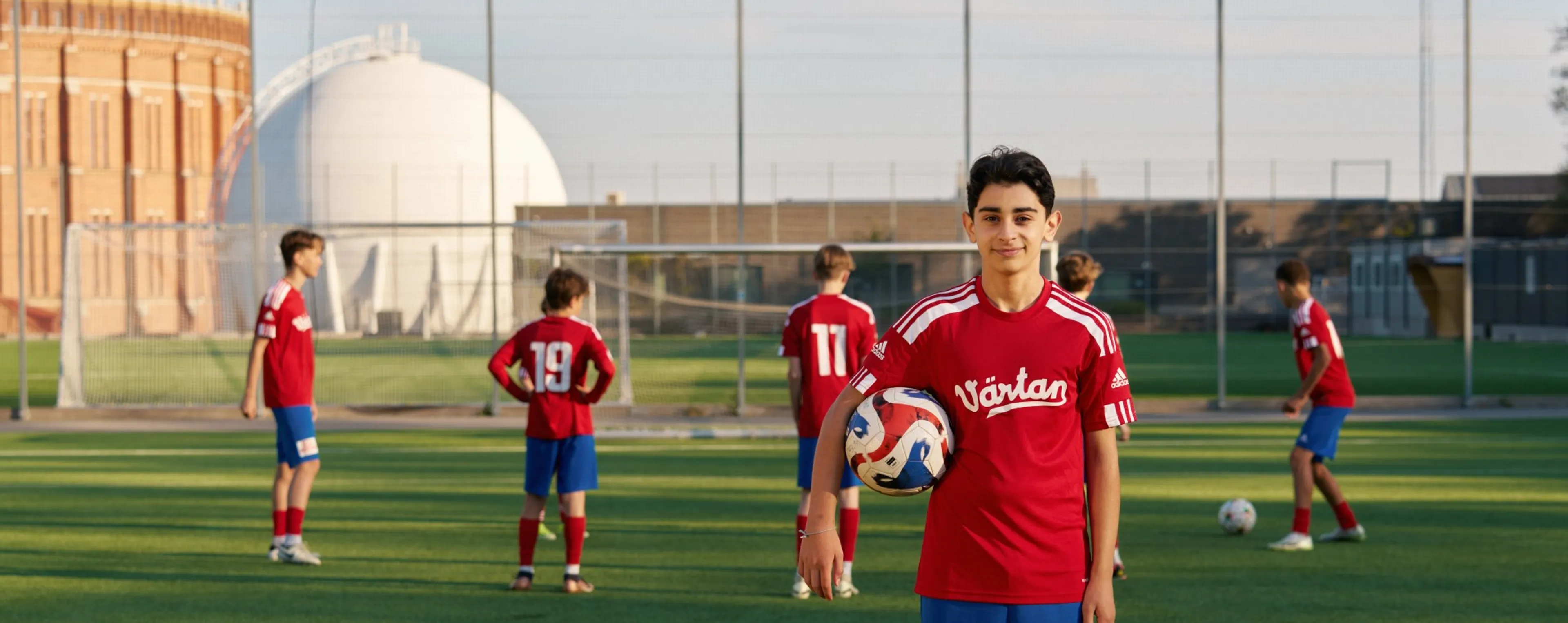 Ett kille i fotbollskläder som har en fotboll under armen och sitt lag i bakgrunden