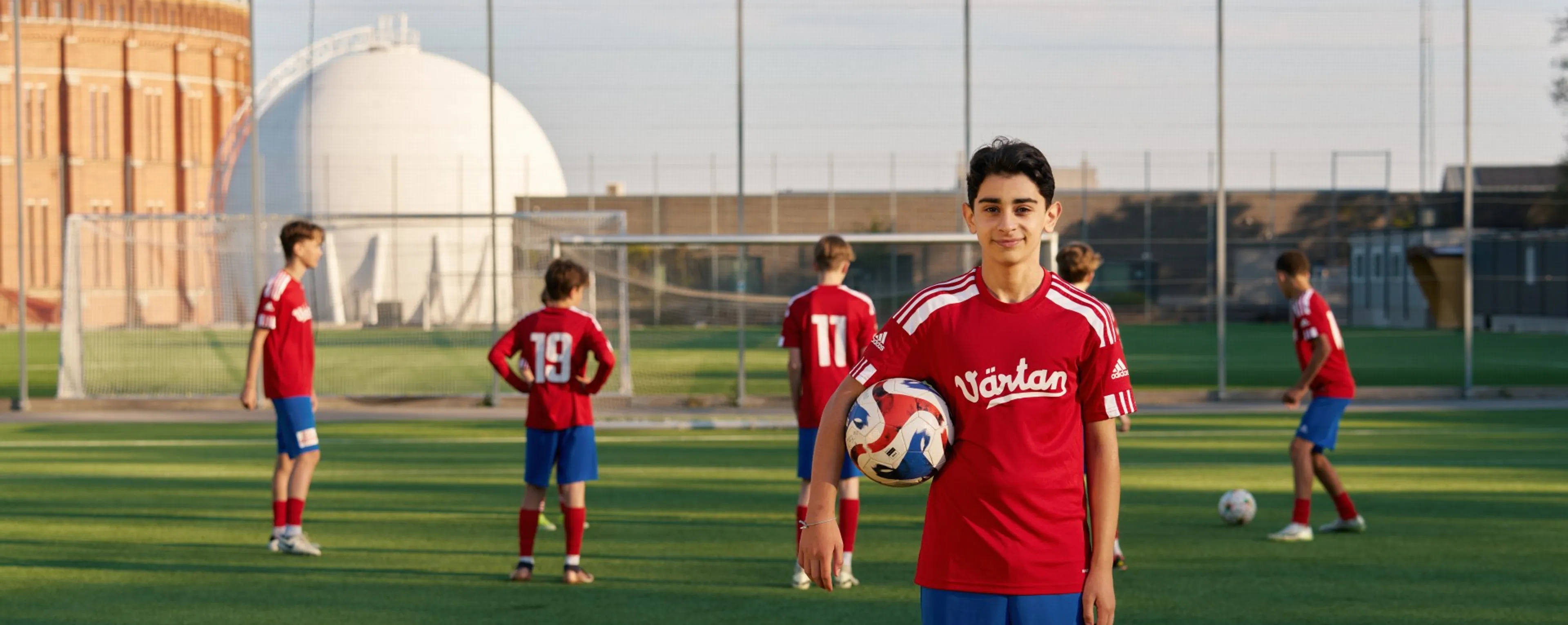 Ett kille i fotbollskläder som har en fotboll under armen och sitt lag i bakgrunden