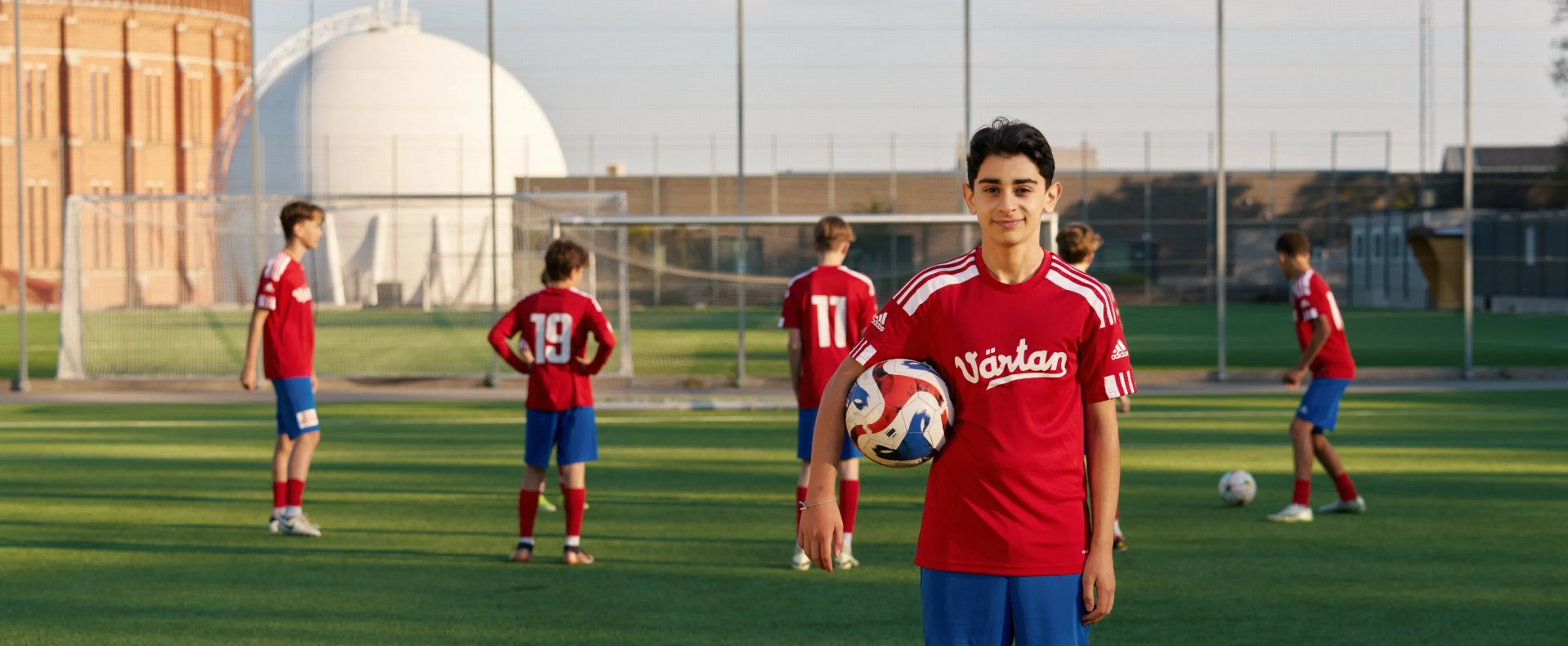 Ett kille i fotbollskläder som har en fotboll under armen och sitt lag i bakgrunden
