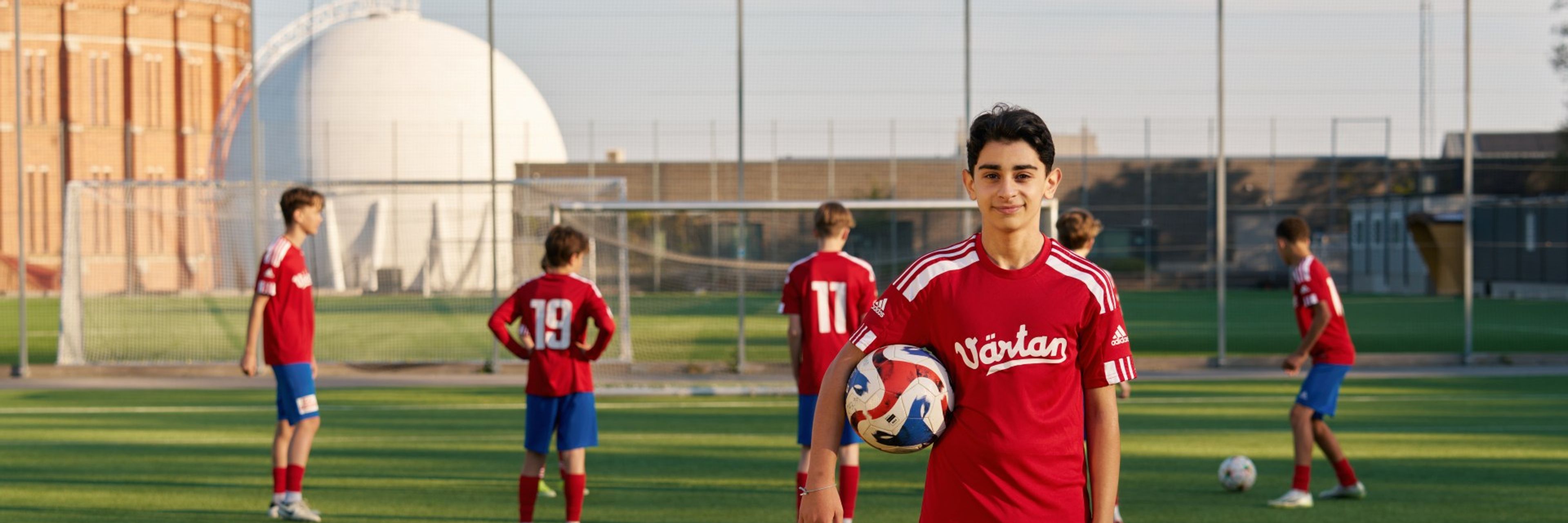Ett kille i fotbollskläder som har en fotboll under armen och sitt lag i bakgrunden