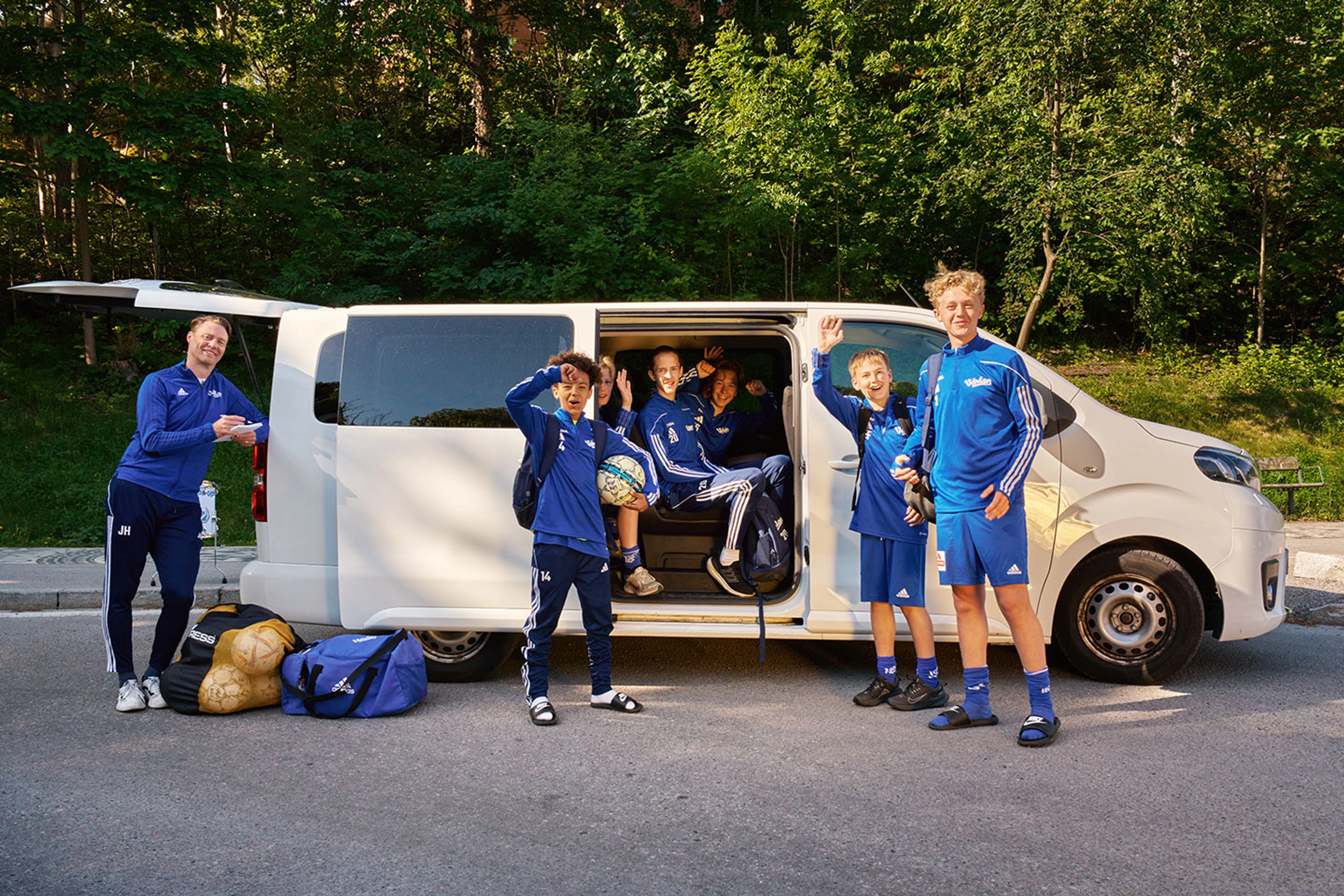 Ett fotbollslag i blå träningskläder står och sitter kring en vit skåpbil med öppna dörrar, redo för träning eller match. Utrustning som väskor och bollar ligger på marken bredvid dem.
