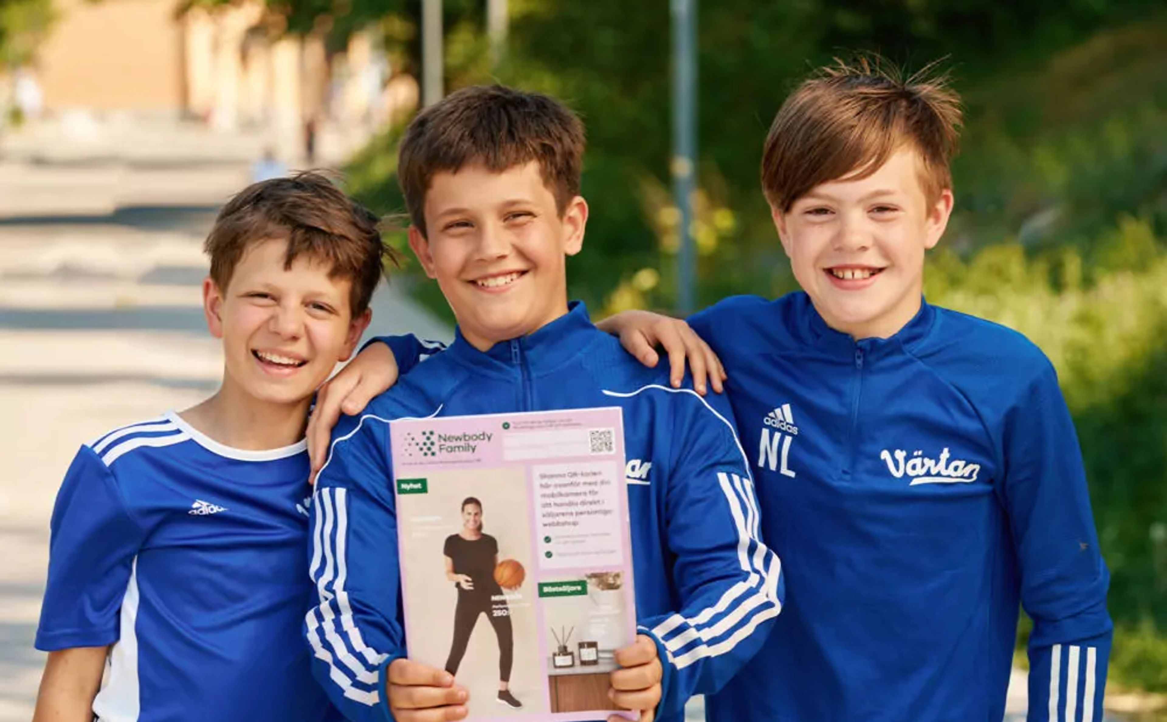 Tre glada pojkar i blå sportkläder står nära varandra utomhus, där en av dem håller upp en katalog med en bild på en person i träningskläder. Bakgrunden visar grönska och en solig dag.