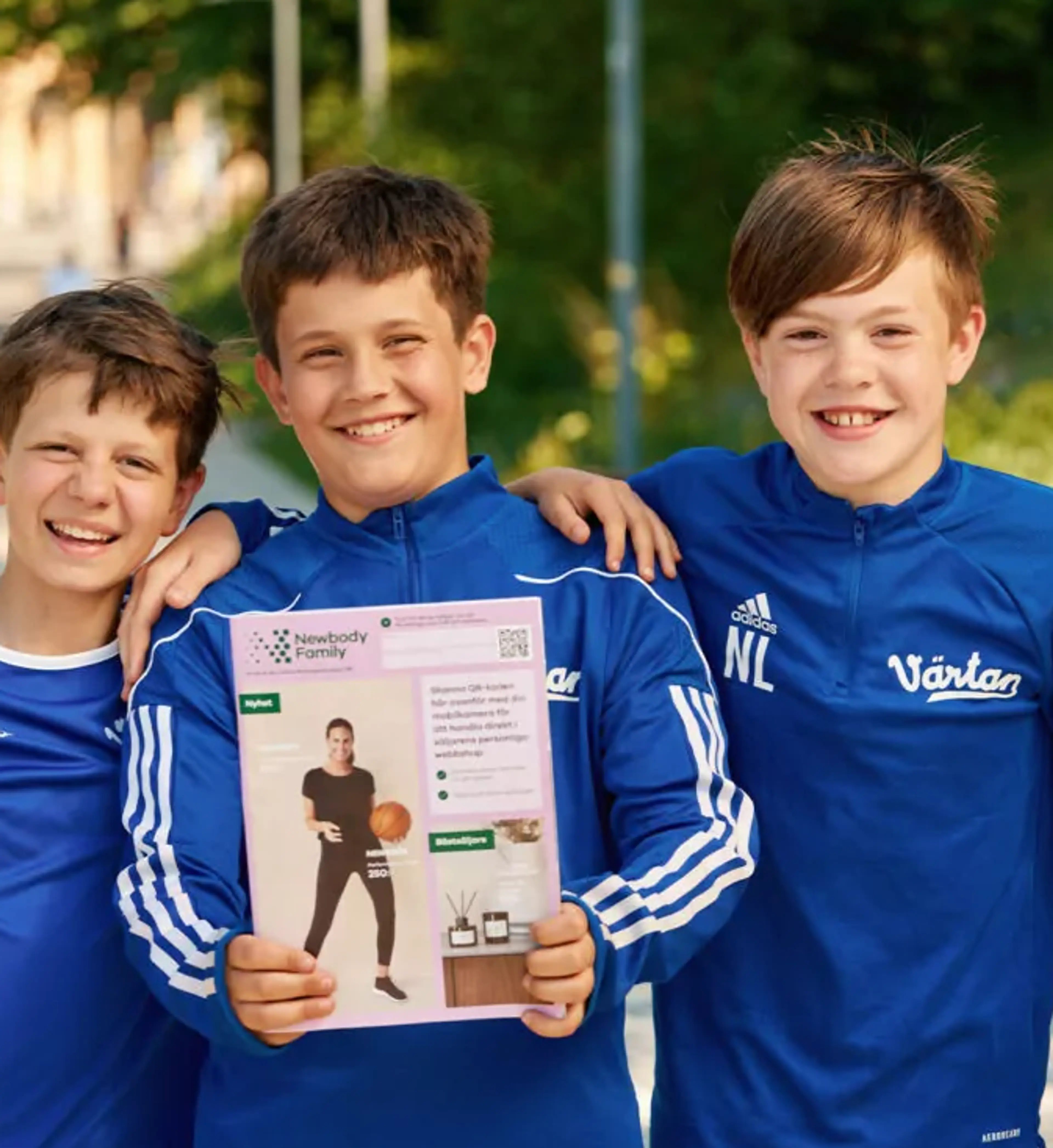 Tre glada pojkar i blå sportkläder står nära varandra utomhus, där en av dem håller upp en katalog med en bild på en person i träningskläder. Bakgrunden visar grönska och en solig dag.