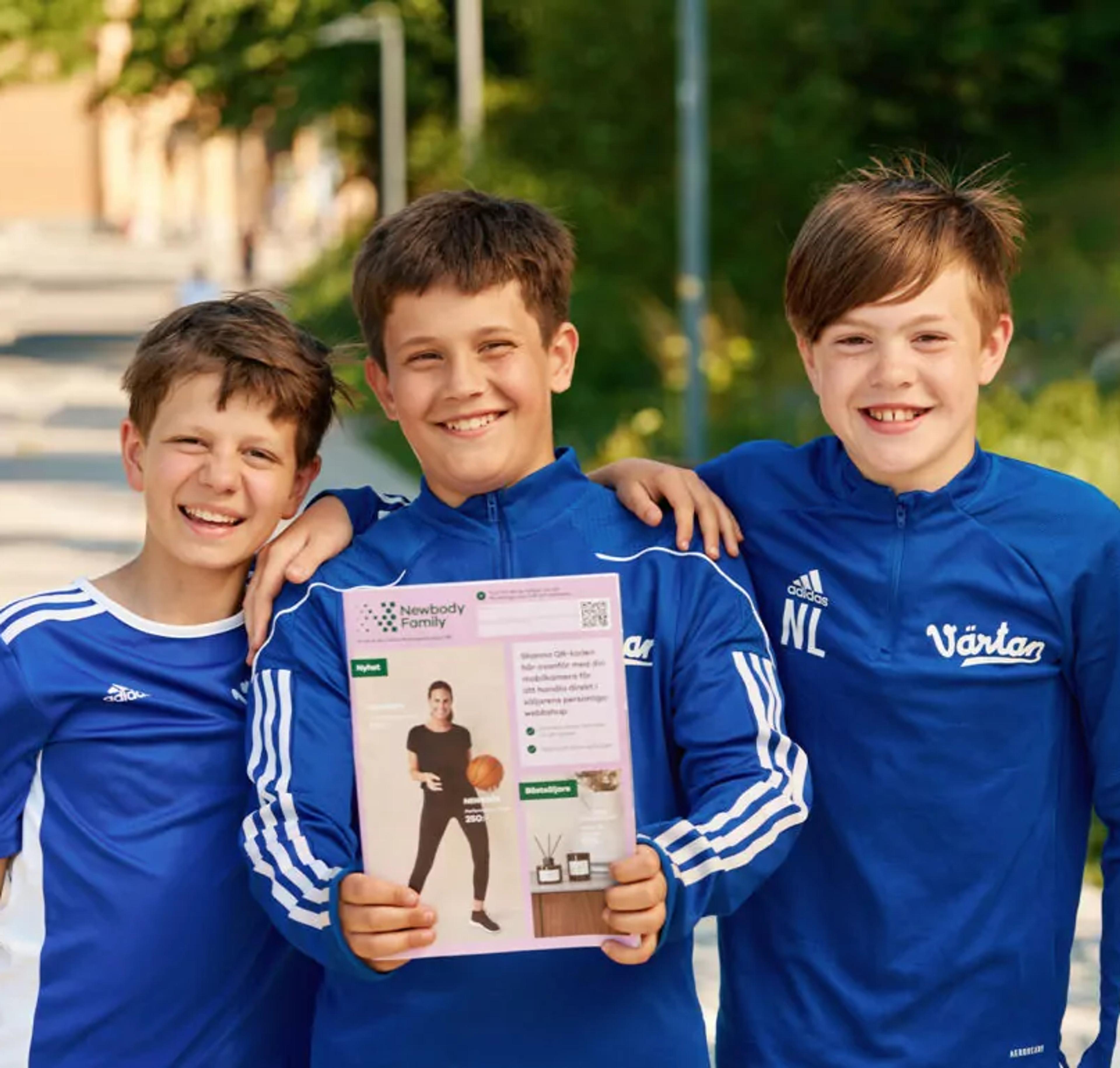 Tre glada pojkar i blå sportkläder står nära varandra utomhus, där en av dem håller upp en katalog med en bild på en person i träningskläder. Bakgrunden visar grönska och en solig dag.