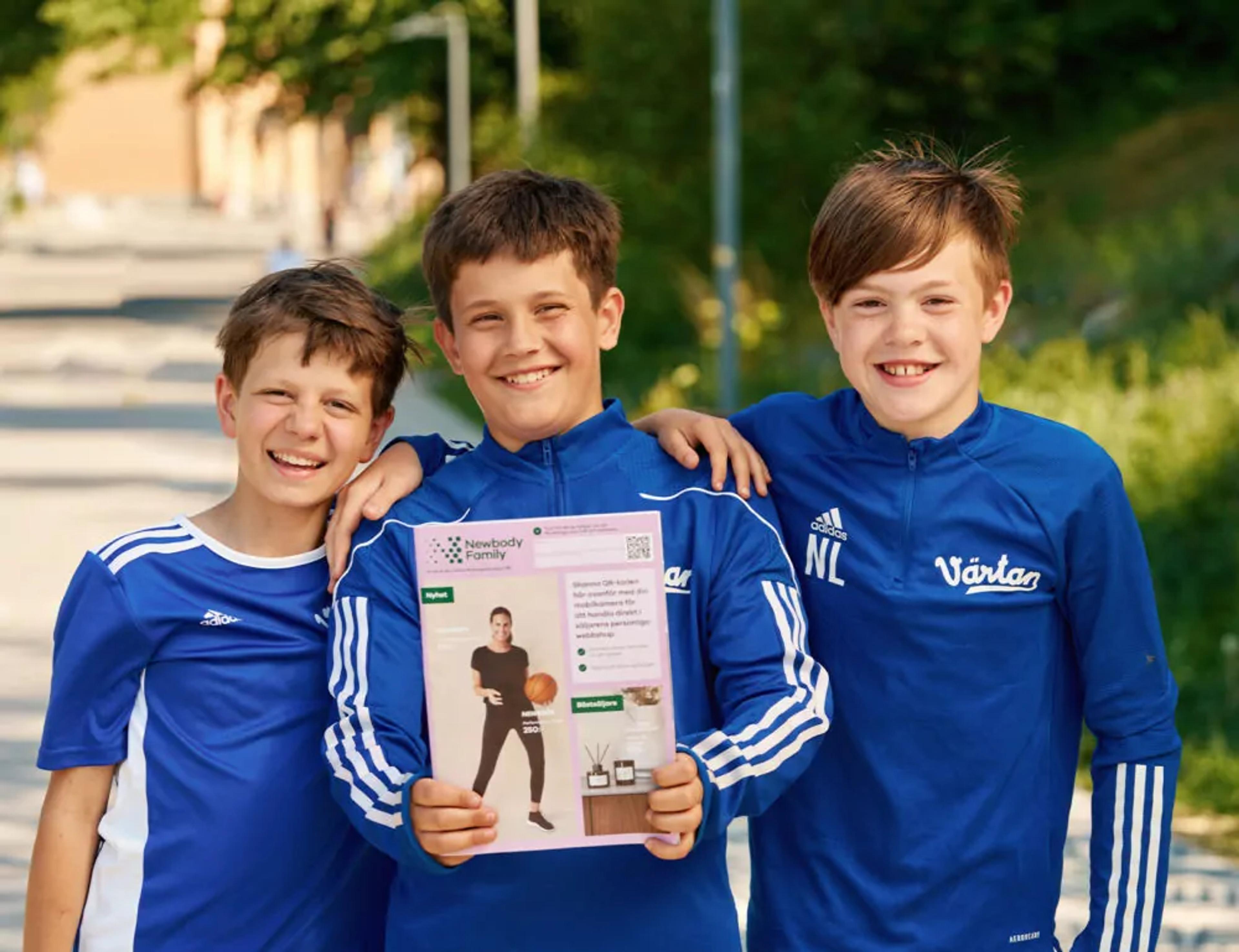 Tre glada pojkar i blå sportkläder står nära varandra utomhus, där en av dem håller upp en katalog med en bild på en person i träningskläder. Bakgrunden visar grönska och en solig dag.