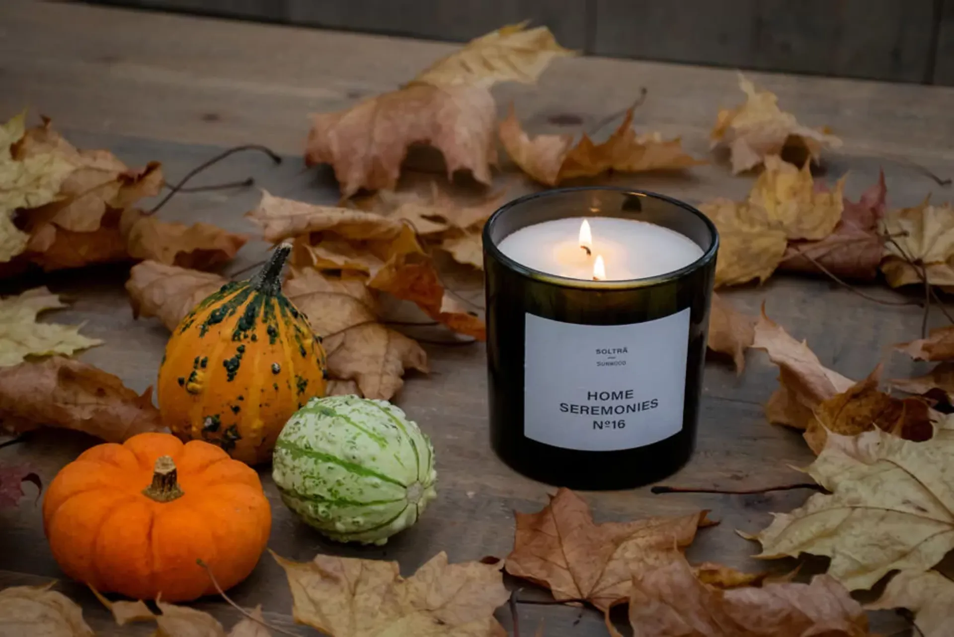 Ett svart glasljus med vit etikett och två brinnande vaxljus bredvid tre små dekorativa pumpor i orange, grön och fläckig orange. Allt ligger på ett träbord omgivet av torra höstlöv.