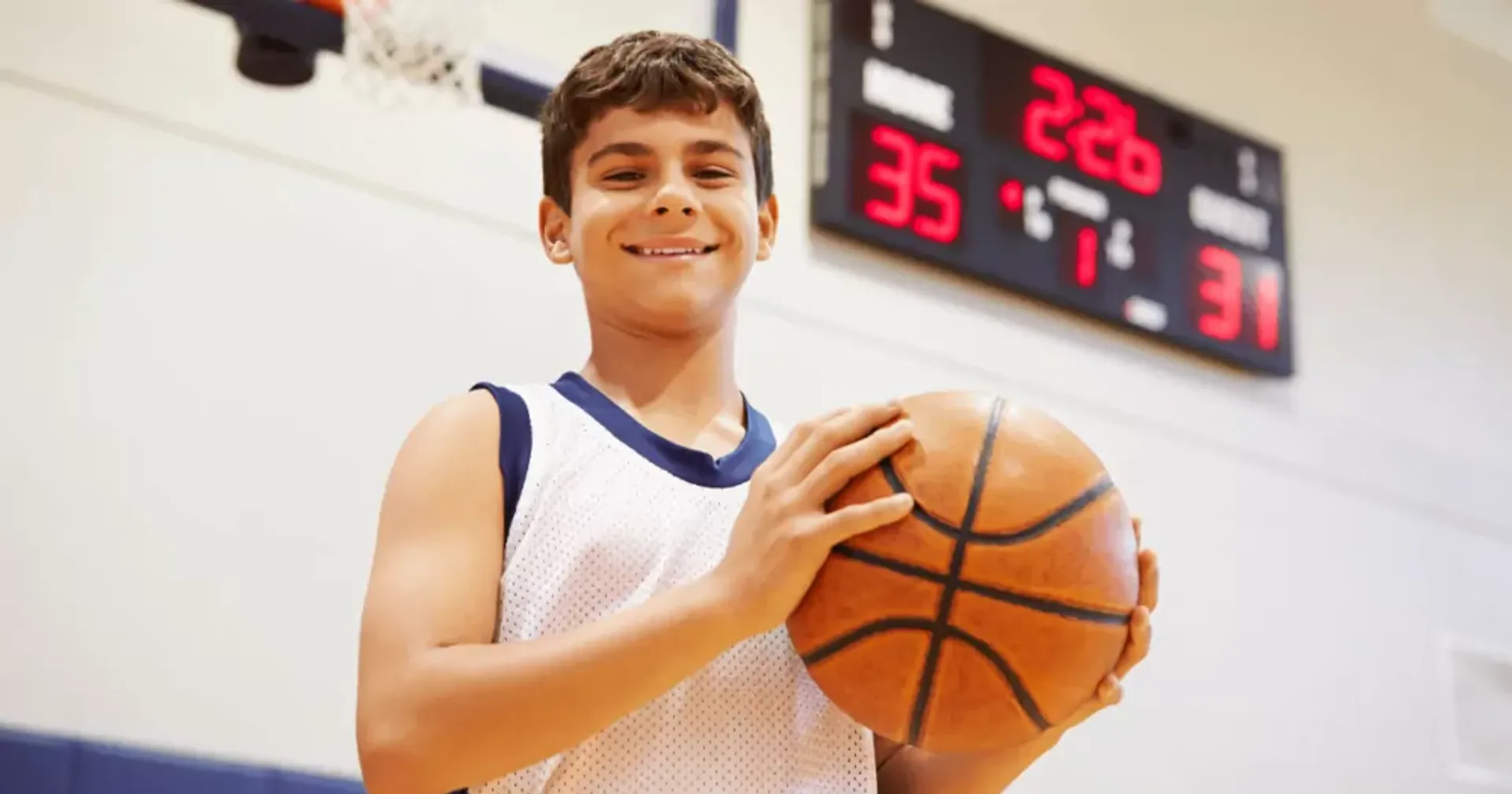 En pojke i vit och blå baskettröja håller en basketboll framför sig i en inomhushall. En poängtavla med siffrorna 35–31 syns i bakgrunden.