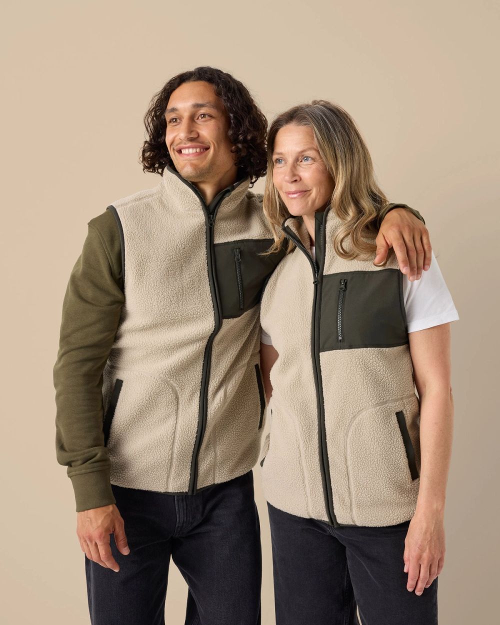 Två personer bär matchande beige fleecevästar med mörkgrönt detaljerat bröstficka och dragkedjor. Västarna är mjuka och värmande med en hög krage.