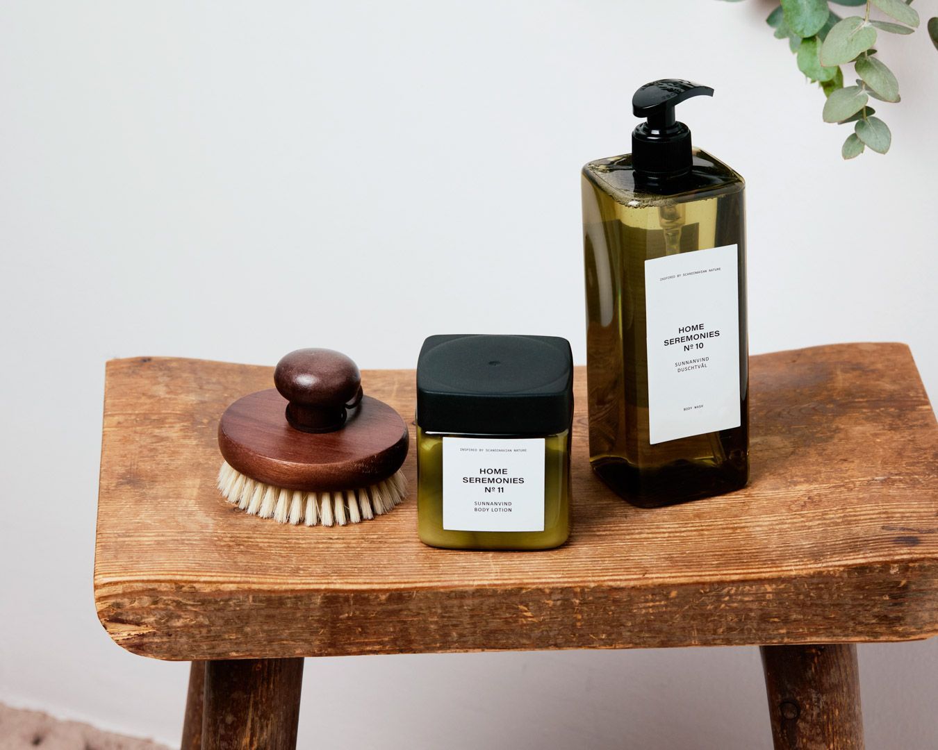 Ett set för kroppsvård med en naturlig kroppsskrubbborste, en burk med kroppslotion och en pumpflaska med duschtvål, alla i jordnära färger och enkel design. Produkterna står på en rustik träpall mot en lj