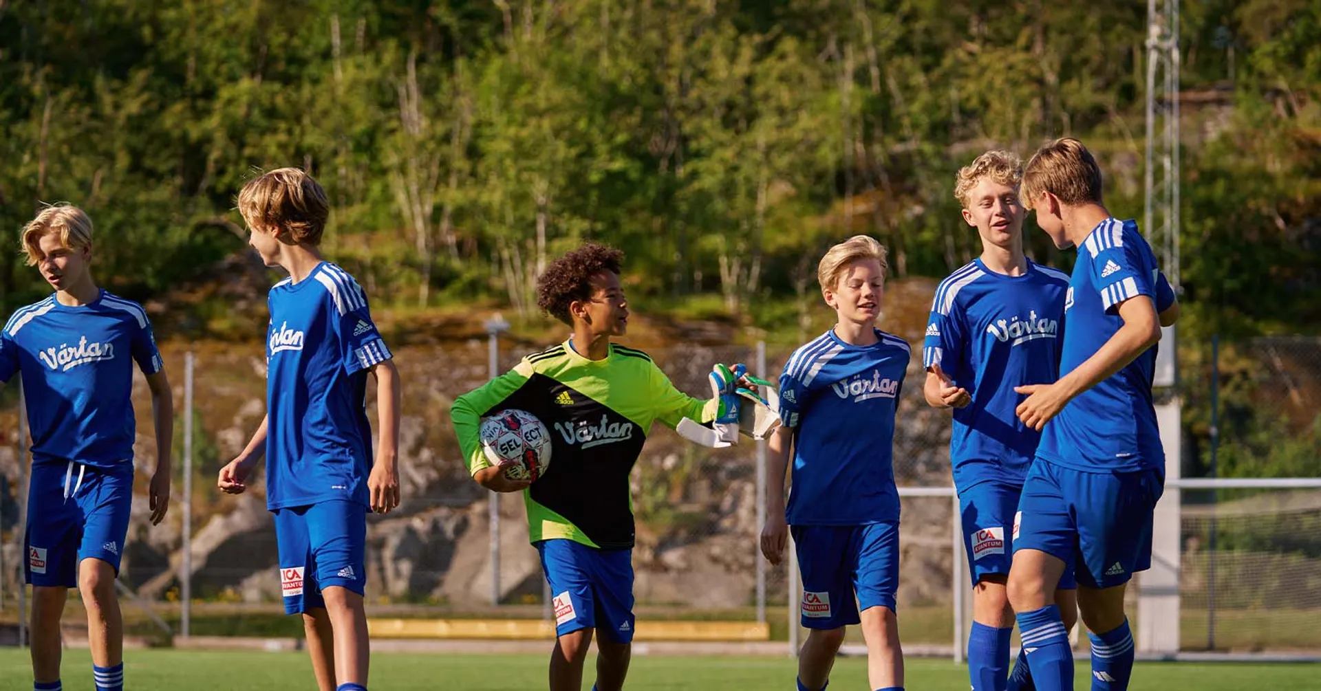 En grupp ungdomsspelare i blå fotbollsdräkter på en utomhusplan, med en målvakt i grön och svart tröja som håller en fotboll. Bakgrunden visar grönska och stenpartier.