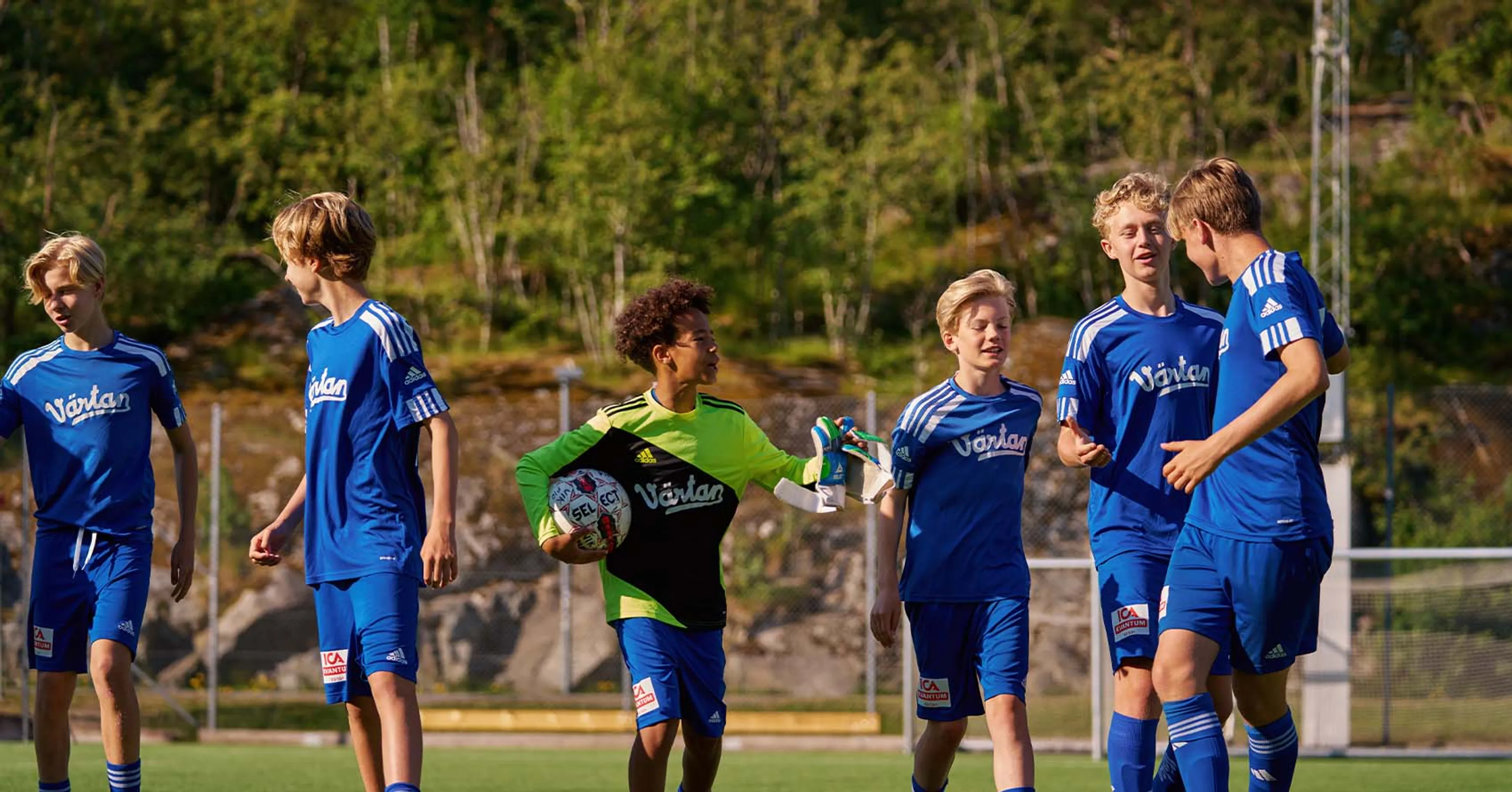 En grupp ungdomsspelare i blå fotbollsdräkter på en utomhusplan, med en målvakt i grön och svart tröja som håller en fotboll. Bakgrunden visar grönska och stenpartier.