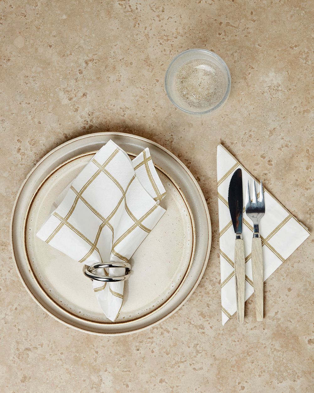 Vita papperservetter med guldglänsande rutmönster, en vikt och placerad i en silverfärgad servettring på två beige tallrikar. En matching servett med kniv och gaffel med ljusa trähandtag ligger intill,