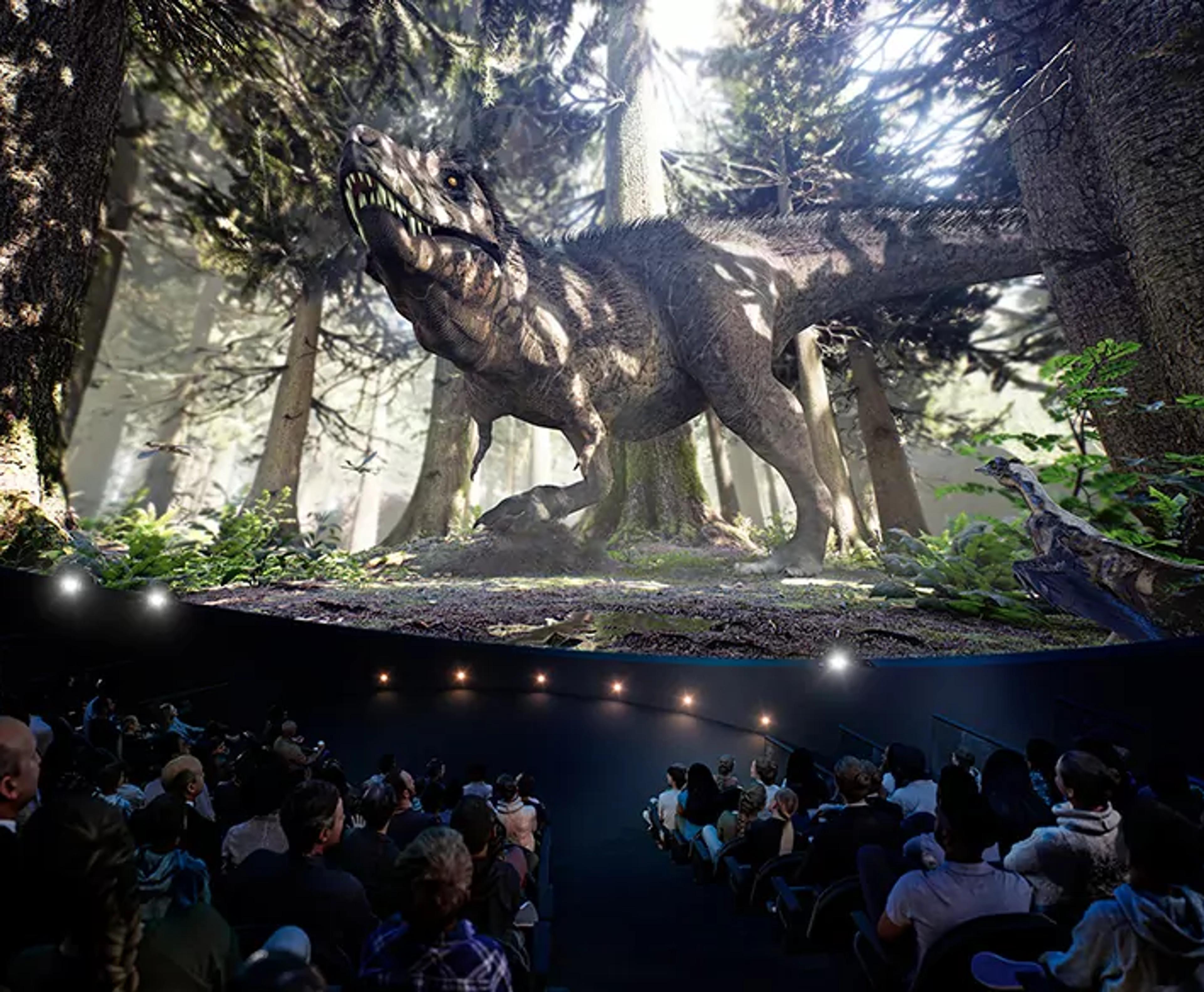 En stor grupp människor sitter tillsammans och tittar på en imponerande, realistisk dinosaurie i en kupolformad bioduk omgiven av skogsmiljö. Scenen skapar en känsla av att befinna sig mitt i en forntida natur.
