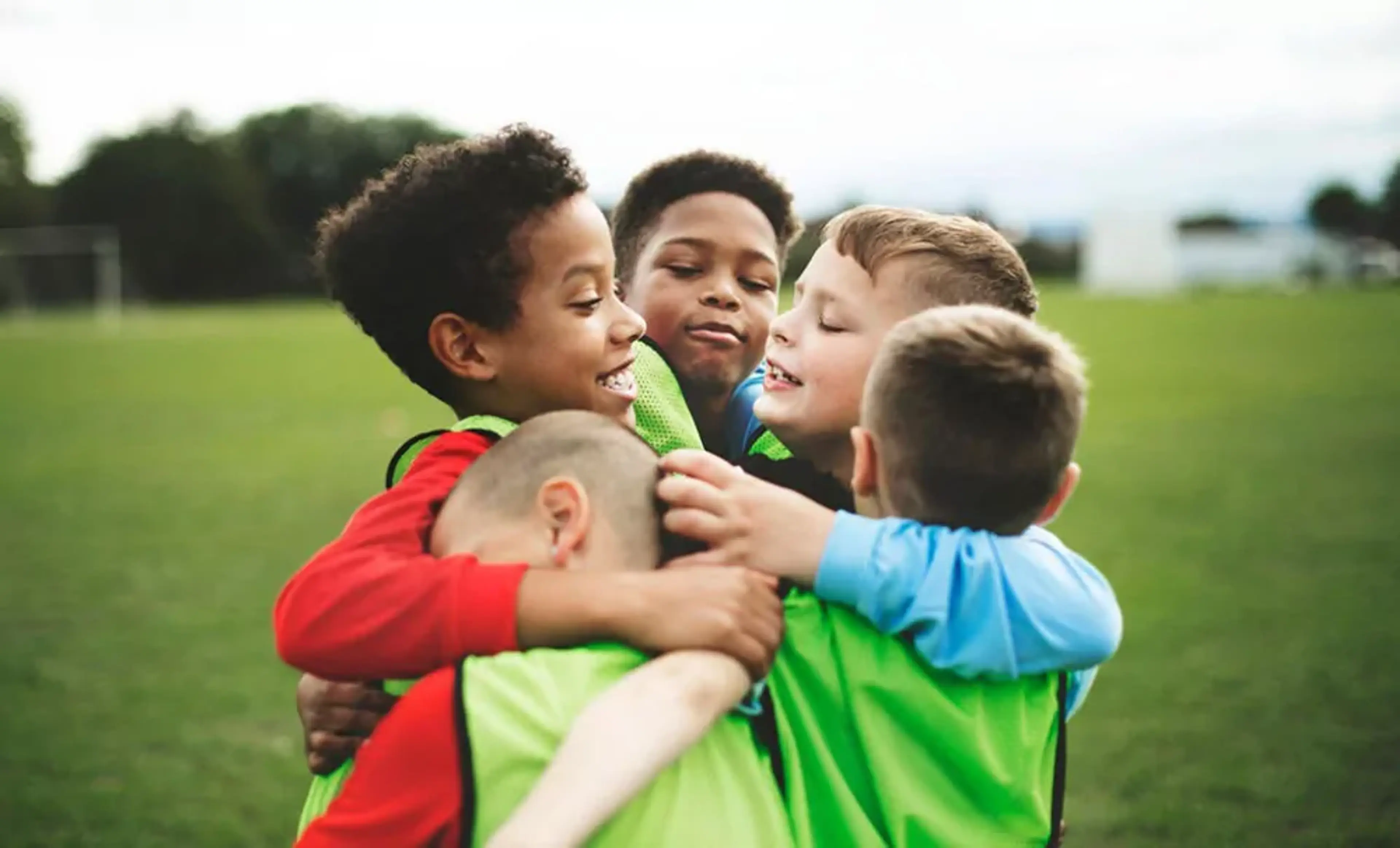 Fyra pojkar i sportkläder kramas tätt på en grön fotbollsplan. De ser glada och sammansvetsade ut efter en match eller träning.