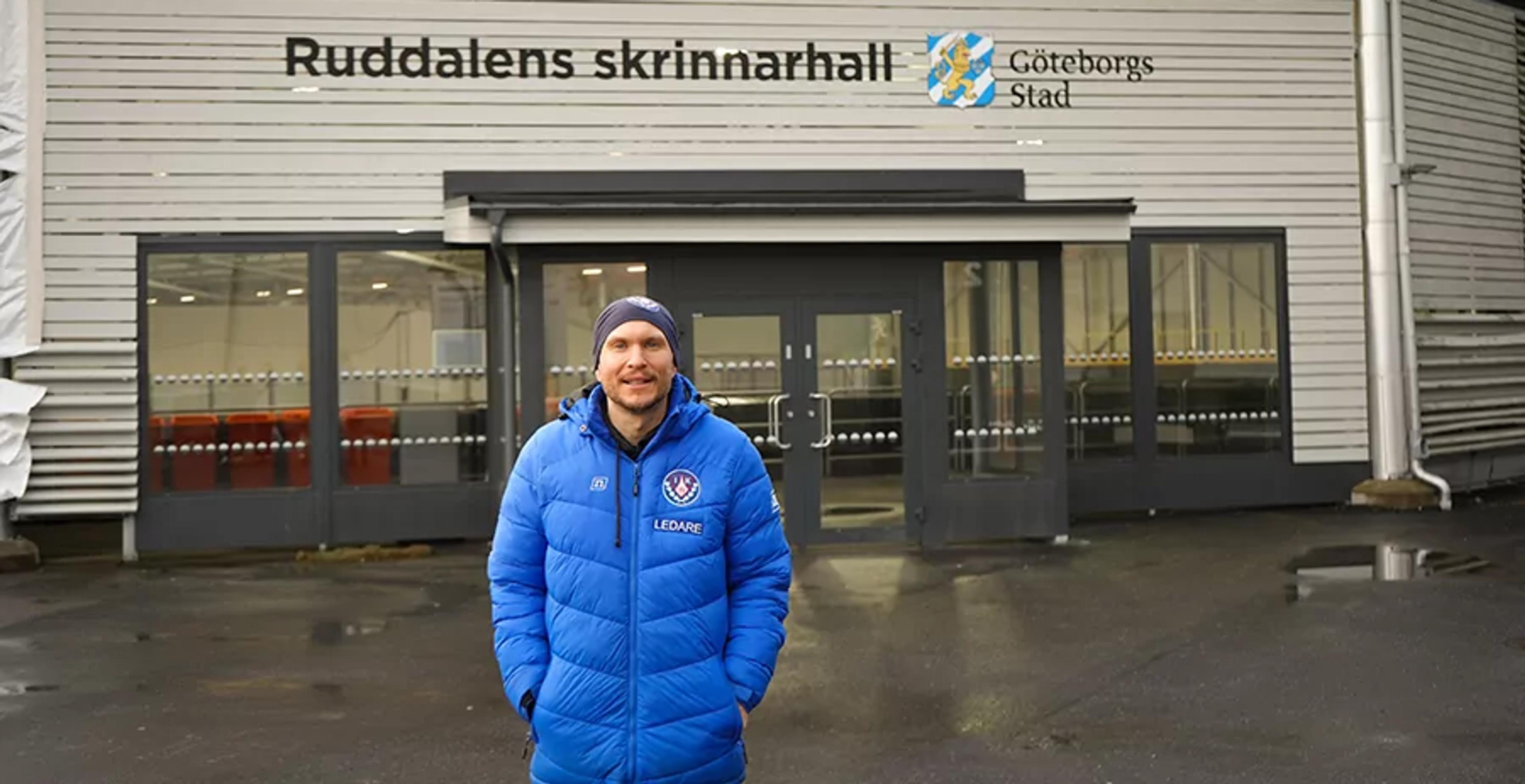 En man i blå jacka med texten "Ledare" står framför entrén till Ruddalens skrinnarhall i Göteborg. Byggnaden är vit och har stora fönster vid ingången.