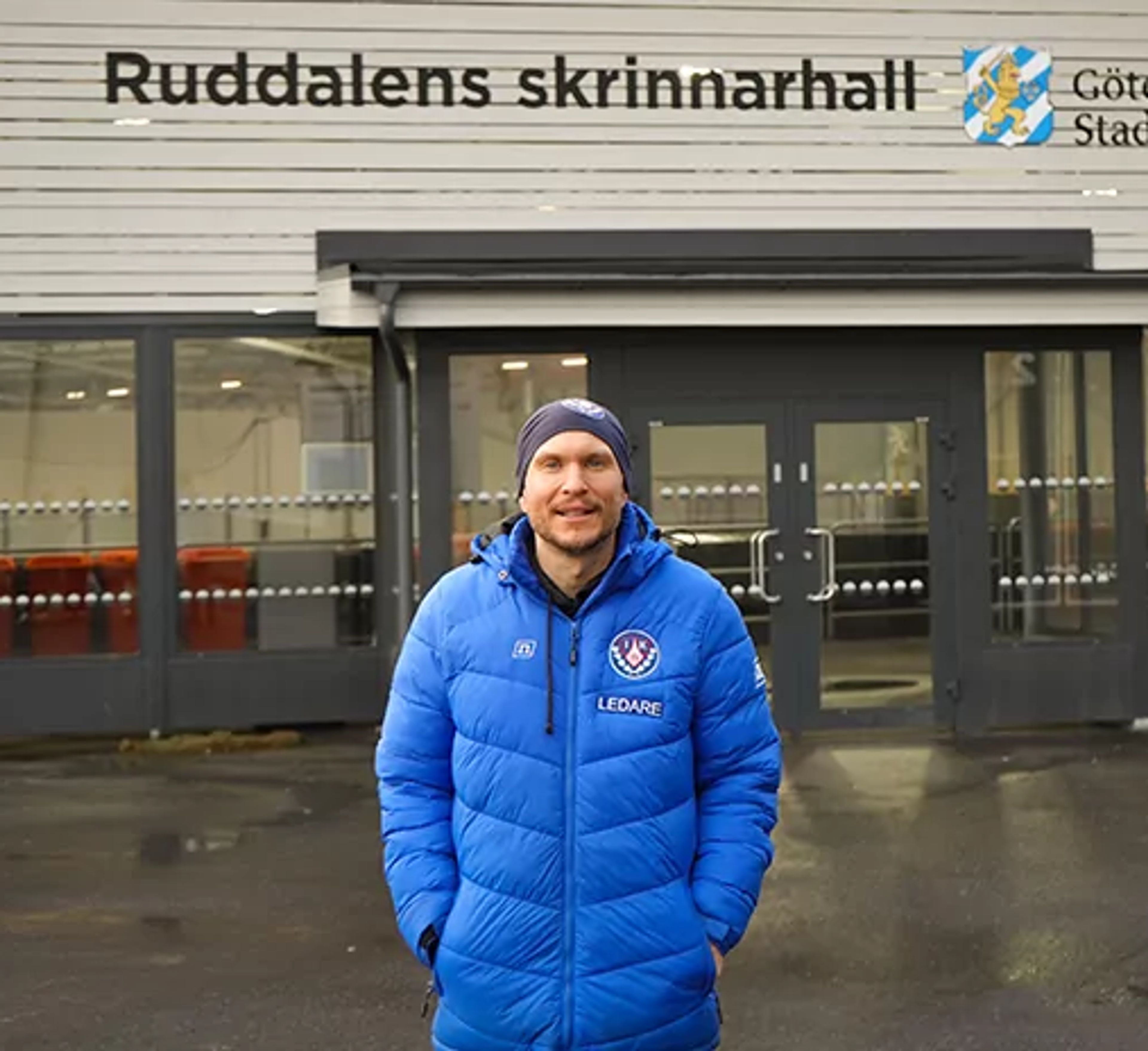 En man i blå jacka med texten "Ledare" står framför entrén till Ruddalens skrinnarhall i Göteborg. Byggnaden är vit och har stora fönster vid ingången.