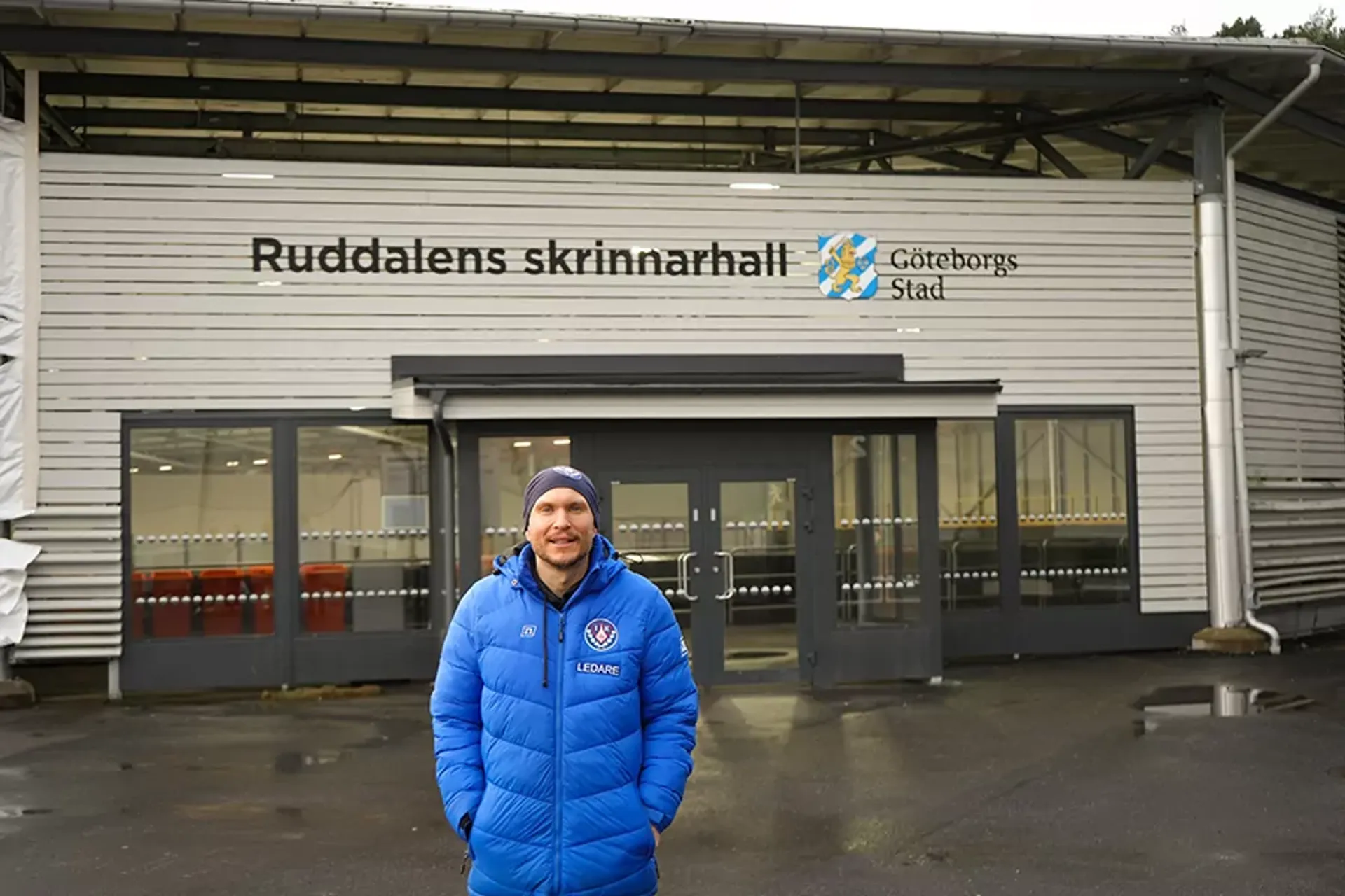 En man i blå jacka med texten "Ledare" står framför entrén till Ruddalens skrinnarhall i Göteborg. Byggnaden är vit och har stora fönster vid ingången.