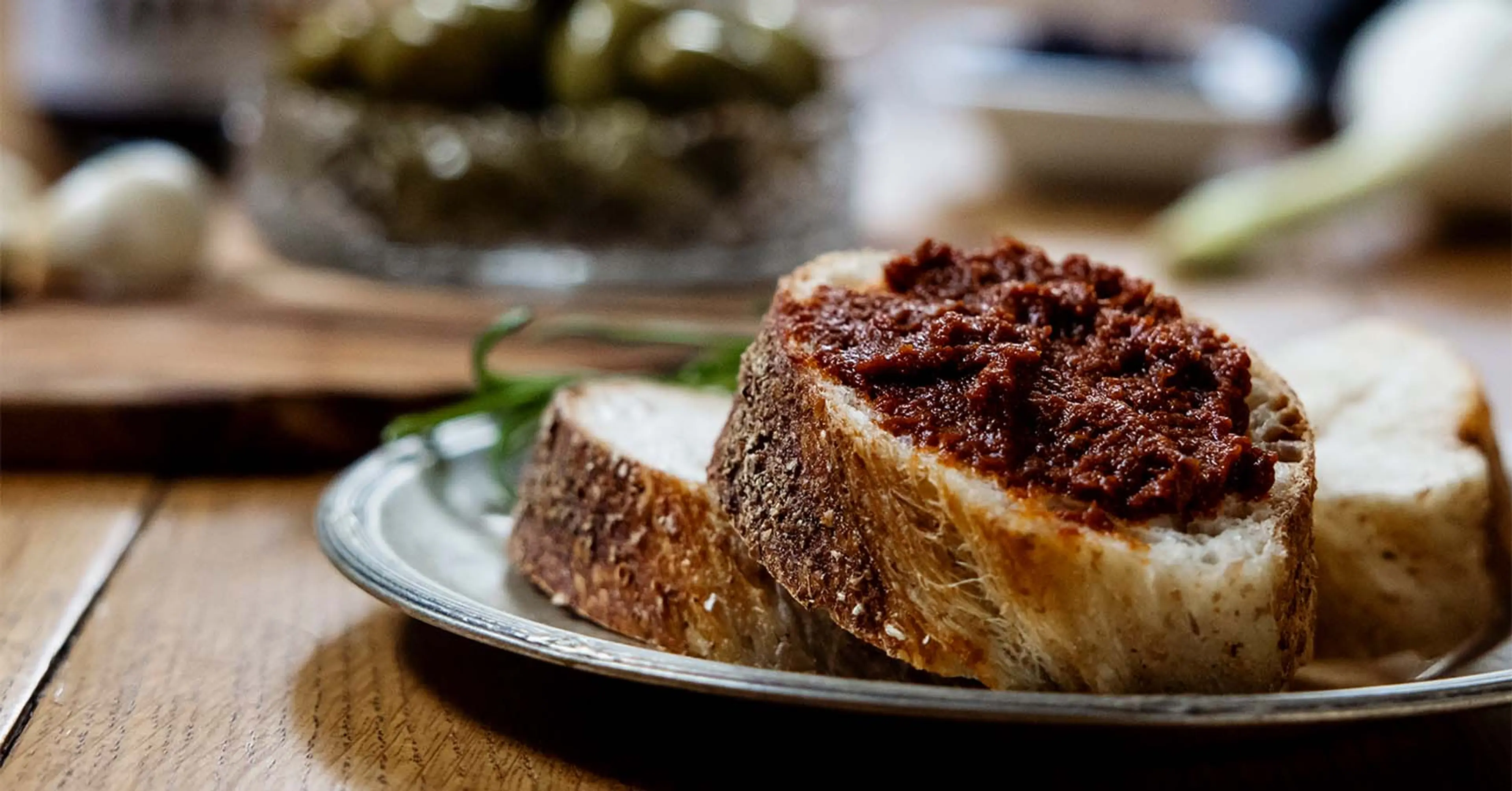 Tre skivor bröd på ett fat, varav en är bred med en krämig röra med djup röd färg. I bakgrunden syns burkar, en skål med gröna oliver och en vitlök.