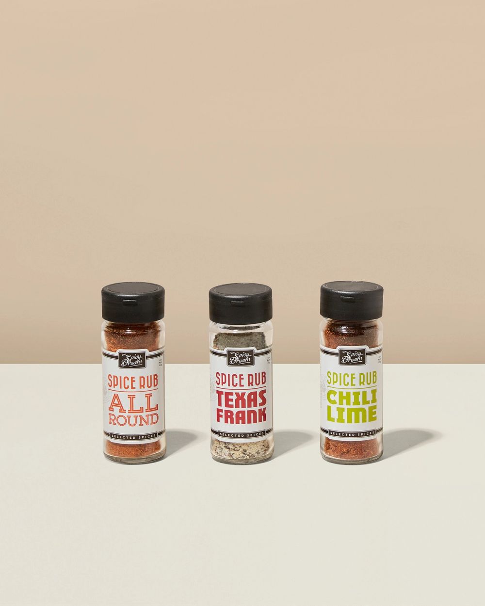 Tre små kryddburkar med svart lock, märkta "All Round", "Texas Frank" och "Chili Lime", fyllda med olika grillkryddor i rött, vitt och grönt.