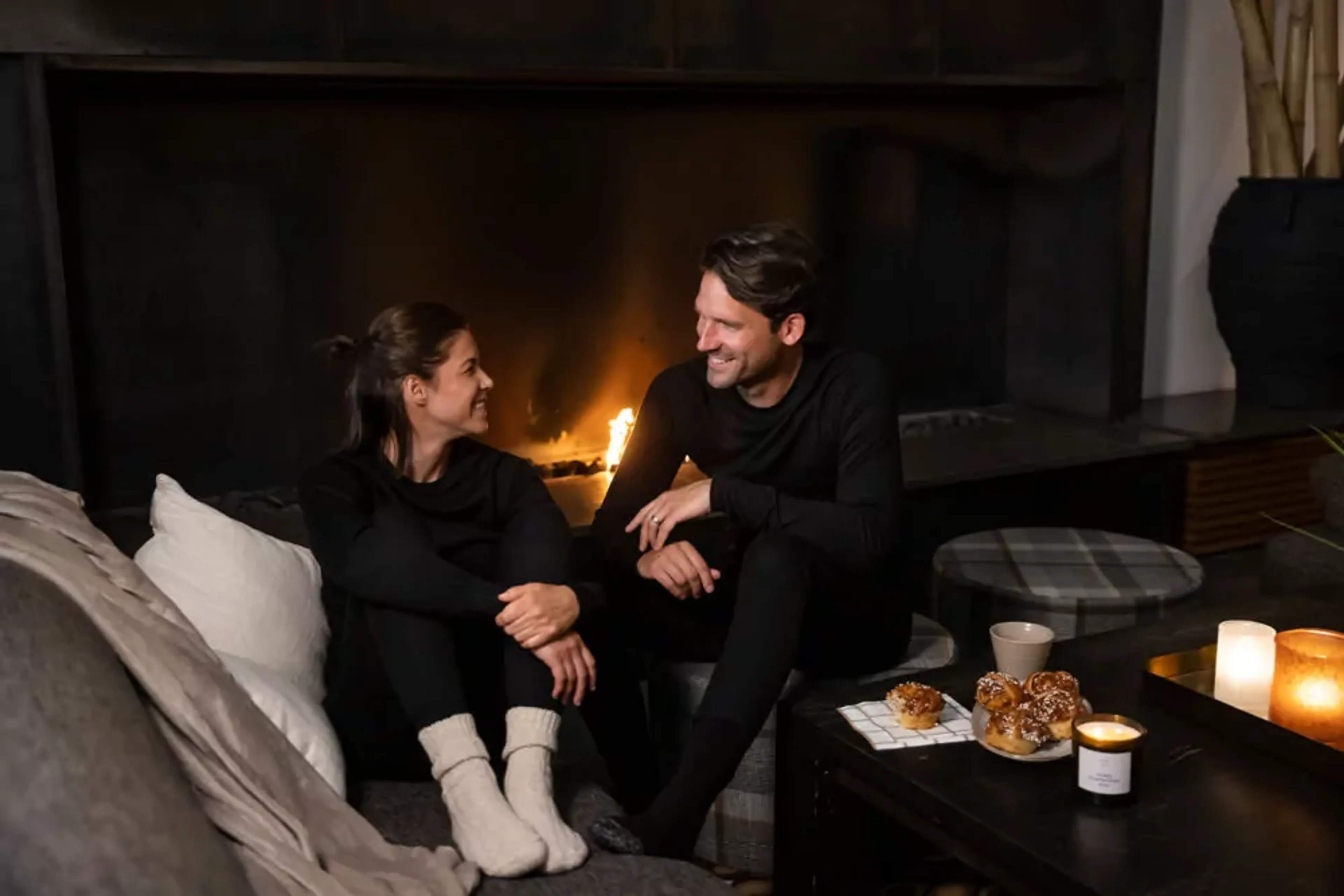 Två personer sitter nära varandra framför en levande brasa i ett mysigt vardagsrum. På bordet framför dem finns bullar, en kopp och tända ljus som sprider ett mjukt sken.