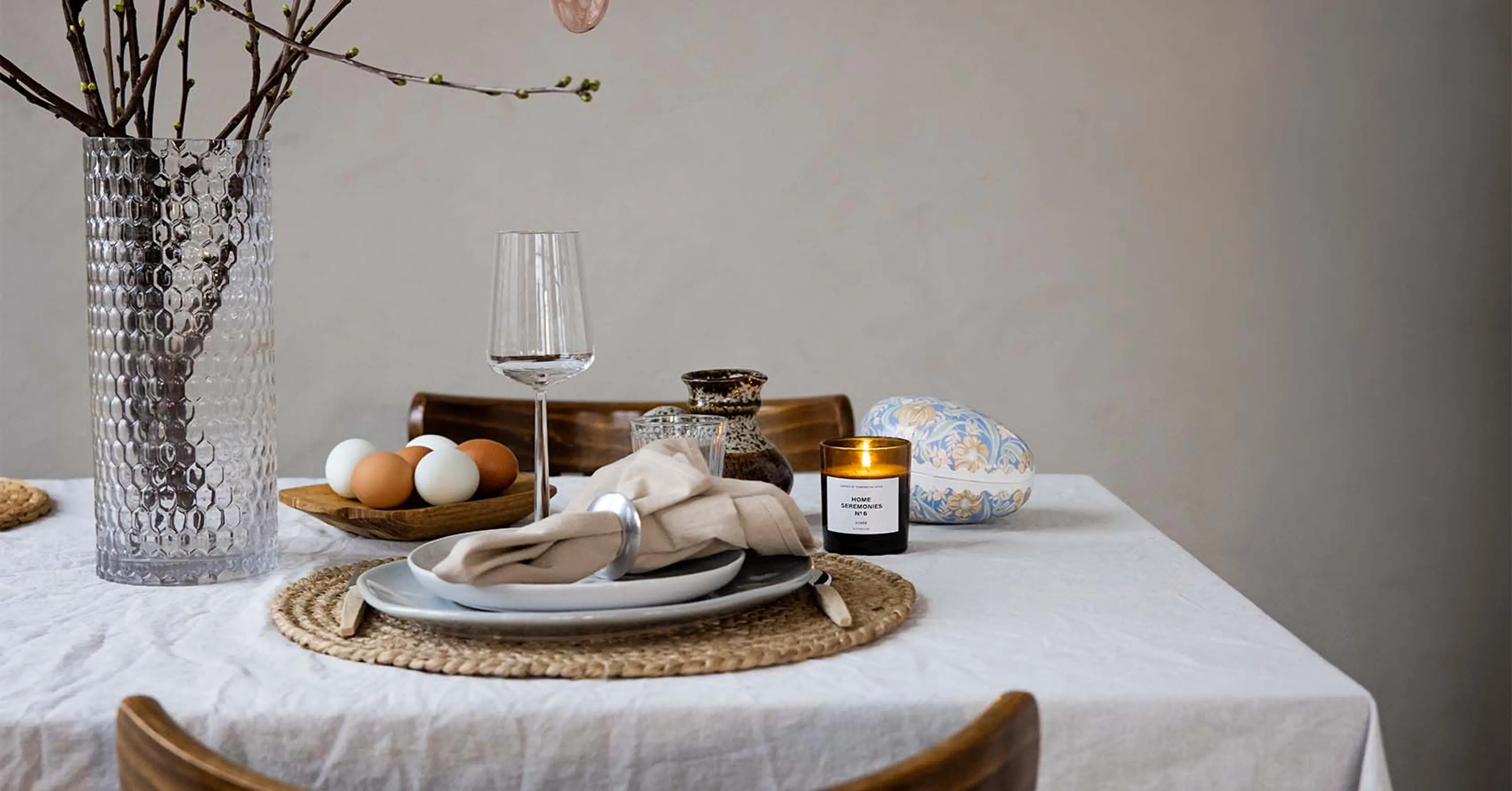 Ett dukat bord med en vit servett i en ring på en ljus tallrik, ett glas, ett blockljus i glas, en skål med vita och bruna ägg samt en vas med kvistar. En vävd rund duklapp ligger under tallriken.