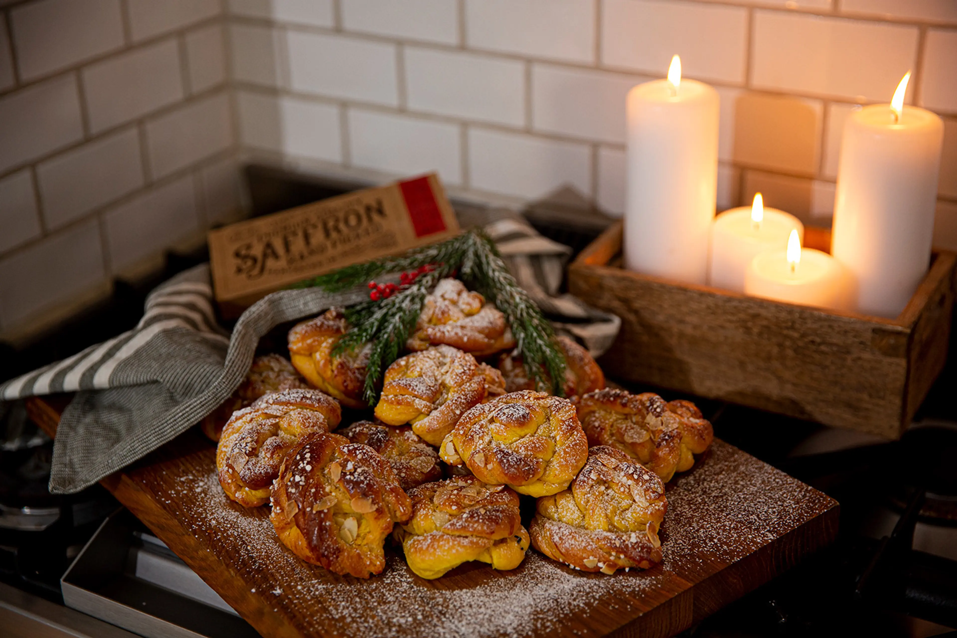 Flera gyllene saffransbullar med pärlsocker och mandelspån ligger på ett träfat, dekorerade med gröna kvistar. I bakgrunden lyser fyra vita ljus i en trälåda.