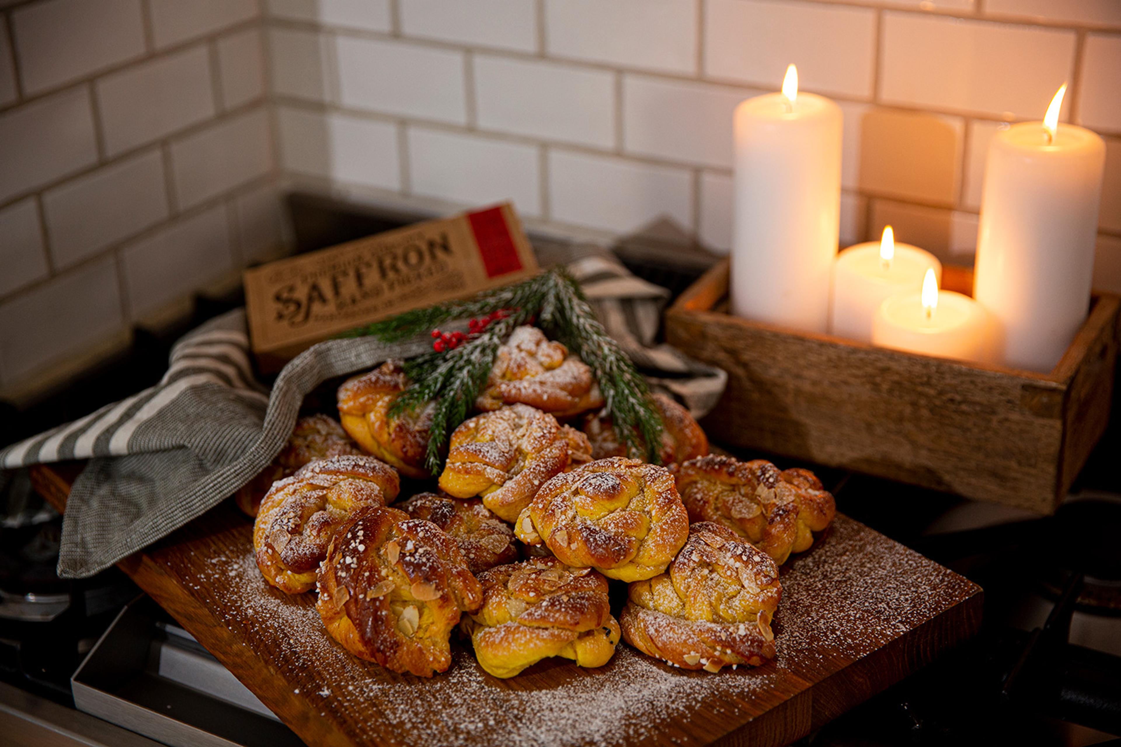 Flera gyllene saffransbullar med pärlsocker och mandelspån ligger på ett träfat, dekorerade med gröna kvistar. I bakgrunden lyser fyra vita ljus i en trälåda.