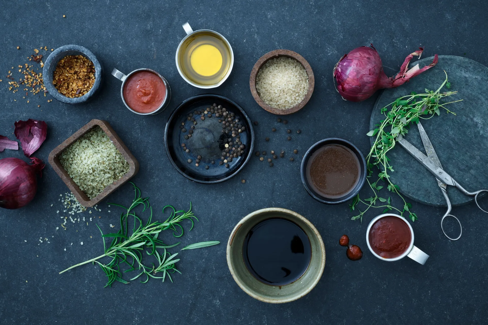 Olika kryddor och örter som svartpeppar, rosmarin, timjan, röd lök samt små skålar med olja, såser och salt ligger prydligt utplacerade på en mörk yta. En sax och ett runt skärbr