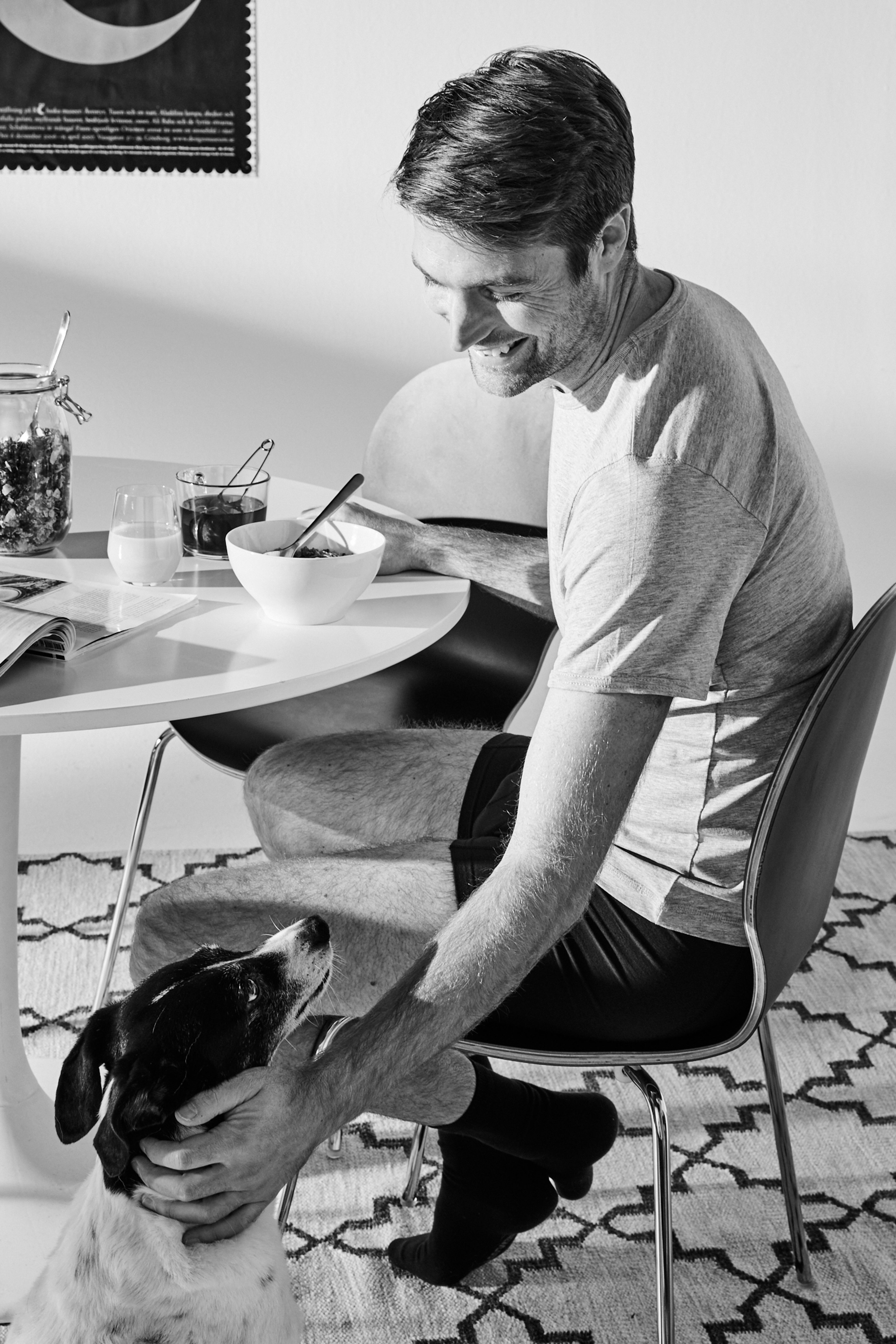 En man i träningskläder sitter vid ett bord med frukost och klappar en hund som tittar upp mot honom. Ett glas mjölk, en skål och burkar står på bordet.