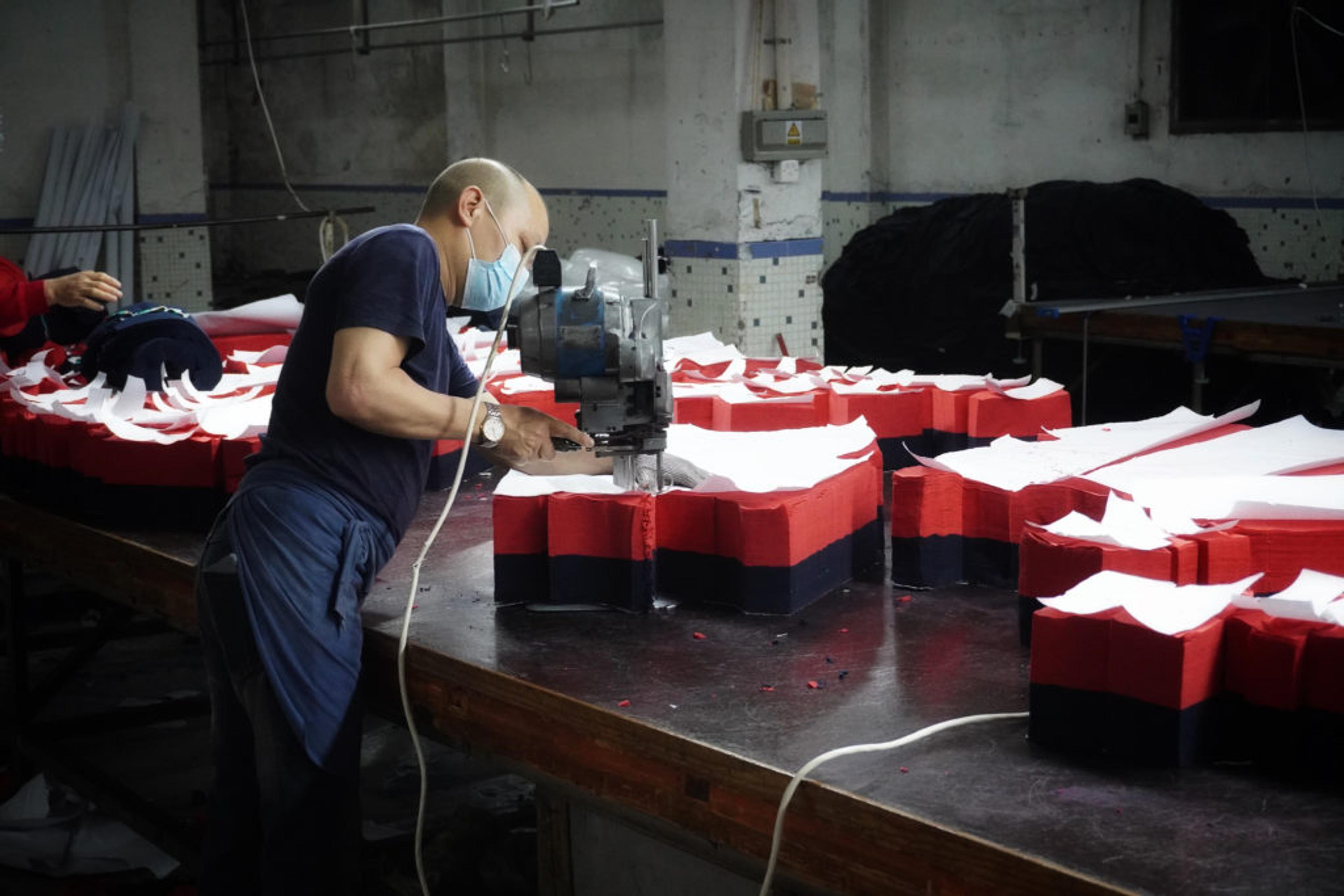 En person med munskydd arbetar med en maskin för att skära eller forma skumgummi i röd, svart och vit färg på ett stort arbetsbord. Industrimiljö med fokus på hantverksmässigt detaljarbete.