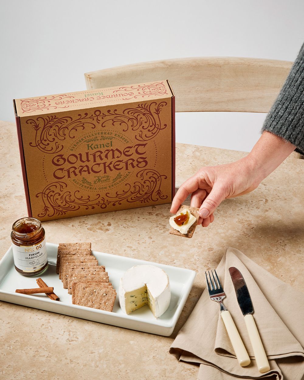 En ask med kanelkryddade gourmetknäckebröd tillsammans med ost, fikonmarmelad och kanelstänger på ett vitt fat. En hand håller upp en knäckebrödsskiva toppad med ost och marmelad.
