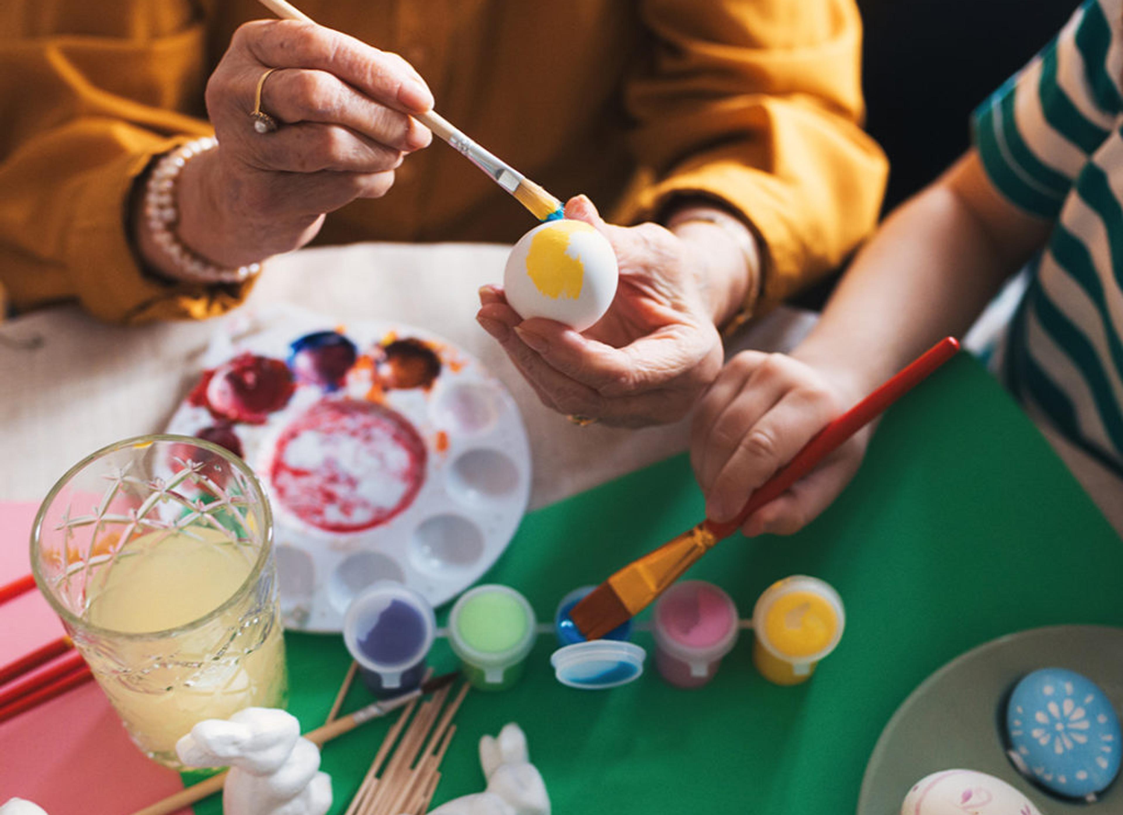 Två personer målar påskägg med färgglada penseldrag, omgivna av små keramikharar och flera färgburkar. Ett fat med färdigmålade ägg och en mugg med ljus dryck står också på bordet.