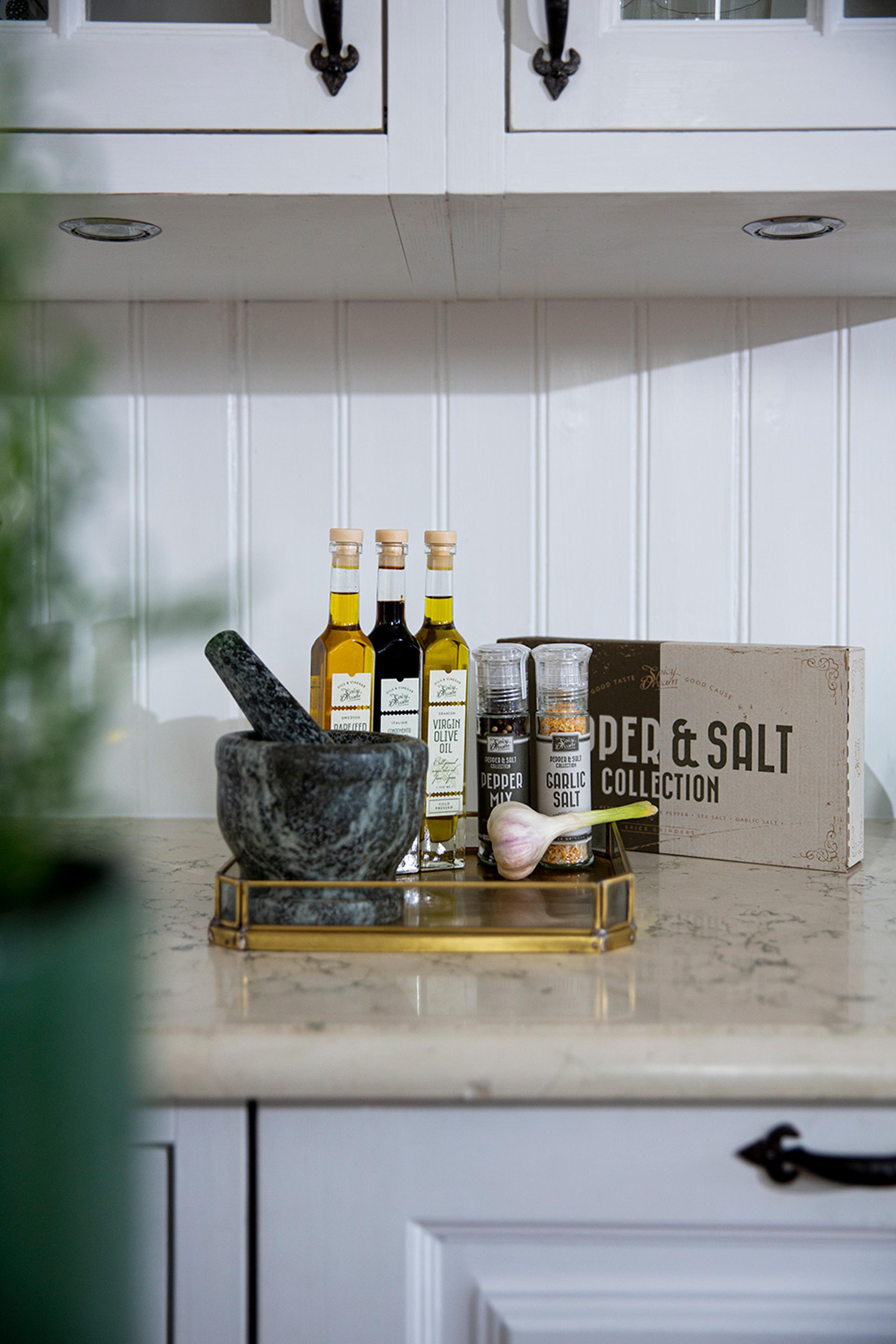 En samling kryddor och oljor i glasflaskor och kryddkvarnar står på en bricka tillsammans med en mortel och en vitlök på ett marmorbord i ett kök. I bakgrunden syns en kartong med texten "PEPPER