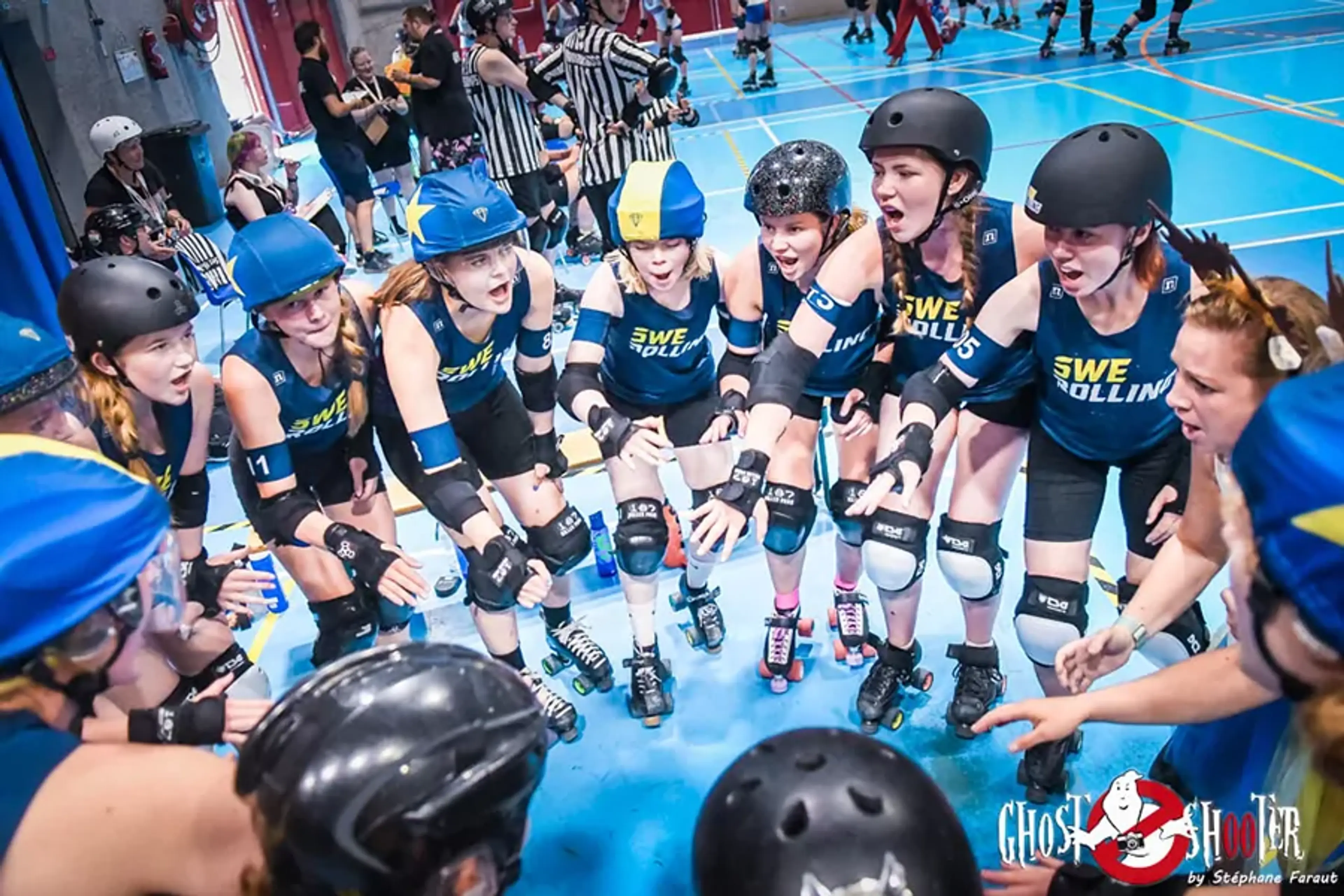 En grupp tjejer i blåa och gula rollerderbyhjälmar och skydd gör en peppande samling på en inomhusbana. Laget har matchkläder med texten "SWE Rolling."