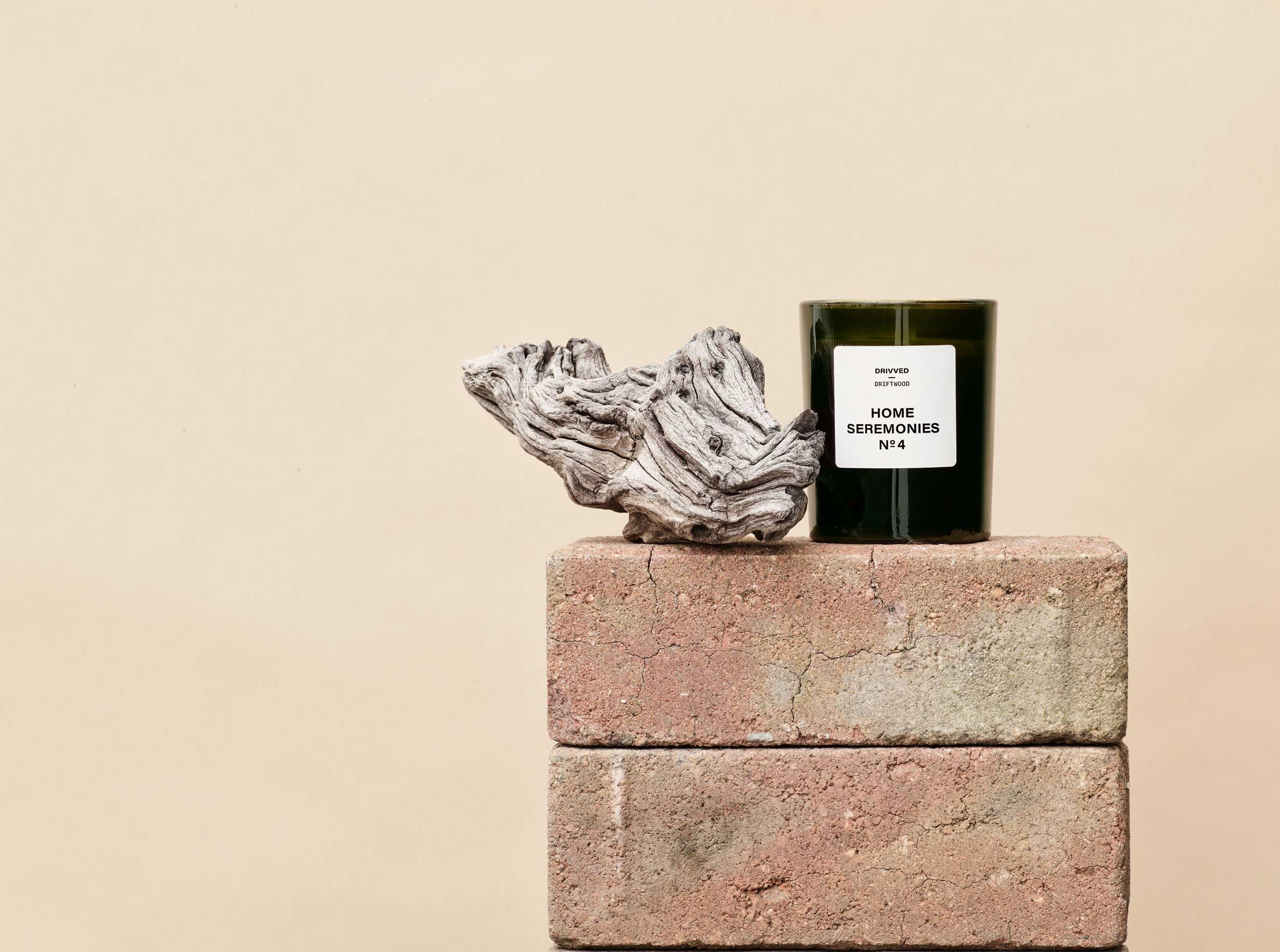 Doftljus i mörkt glas med etikett, placerat på två staplade tegelstenar tillsammans med en bit drivved.