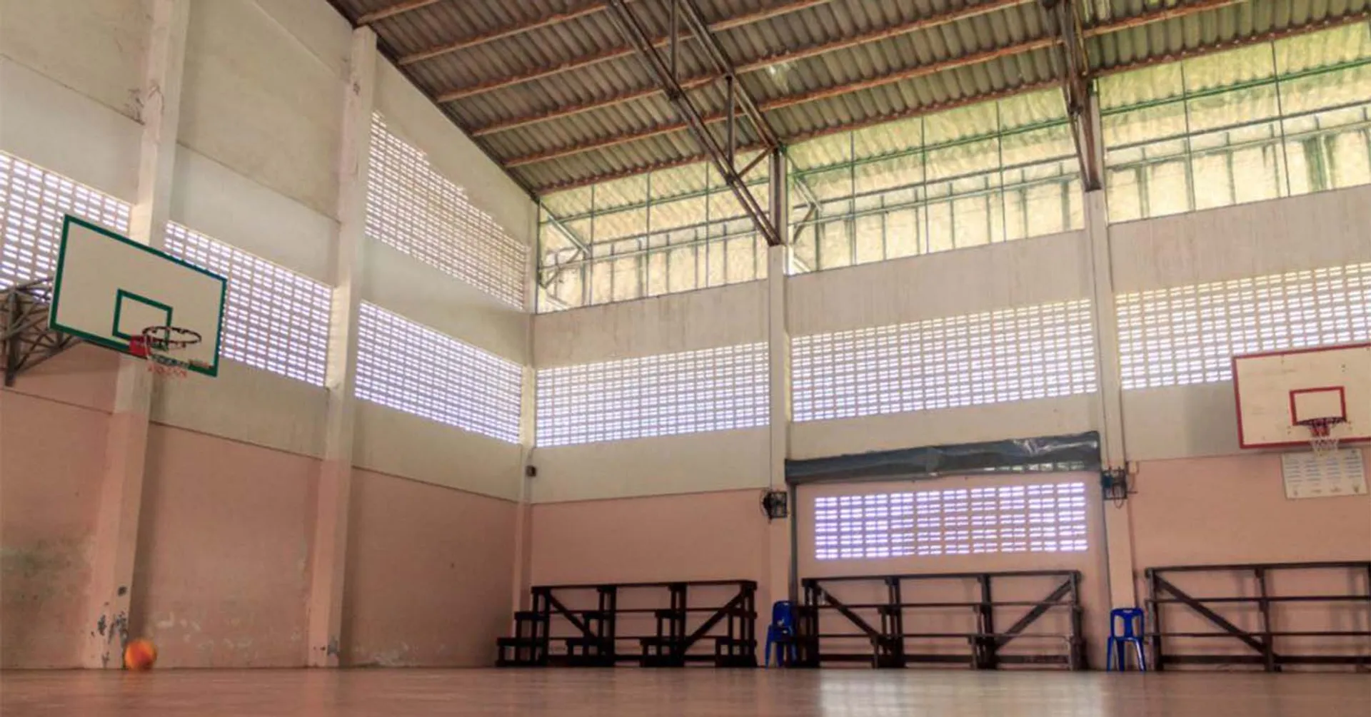 En tom gympasal med högt till tak, två basketkorgar och bänkar längs väggarna. Ljus strömmar in genom höga fönster i väggarna.