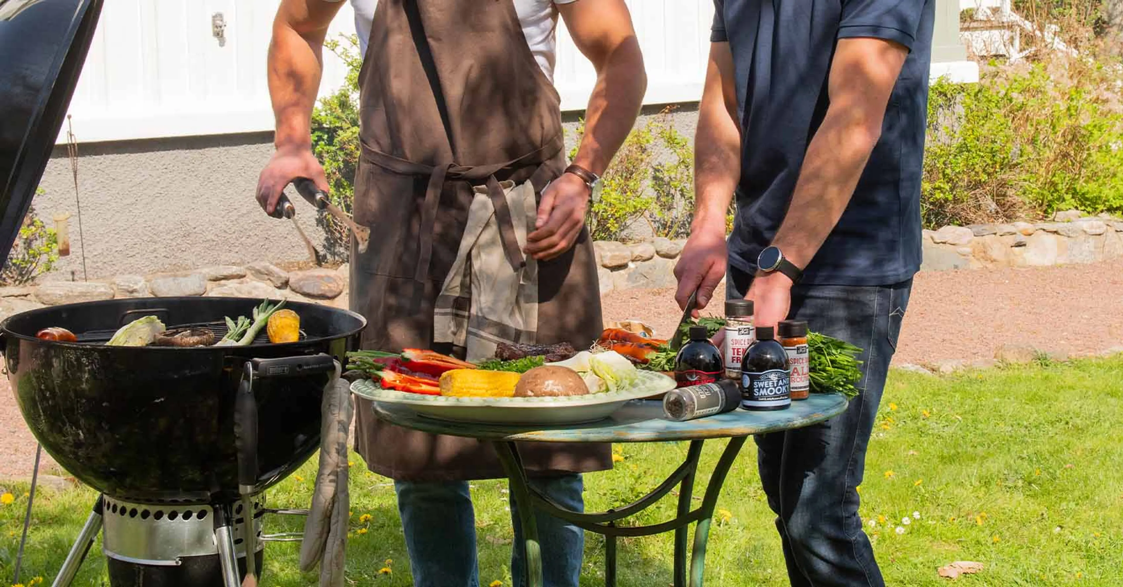 Två personer vid en grill med en tallrik fylld av grönsaker och kryddflaskor på ett bord bredvid. En person håller grilltång och den andra står redo att krydda.