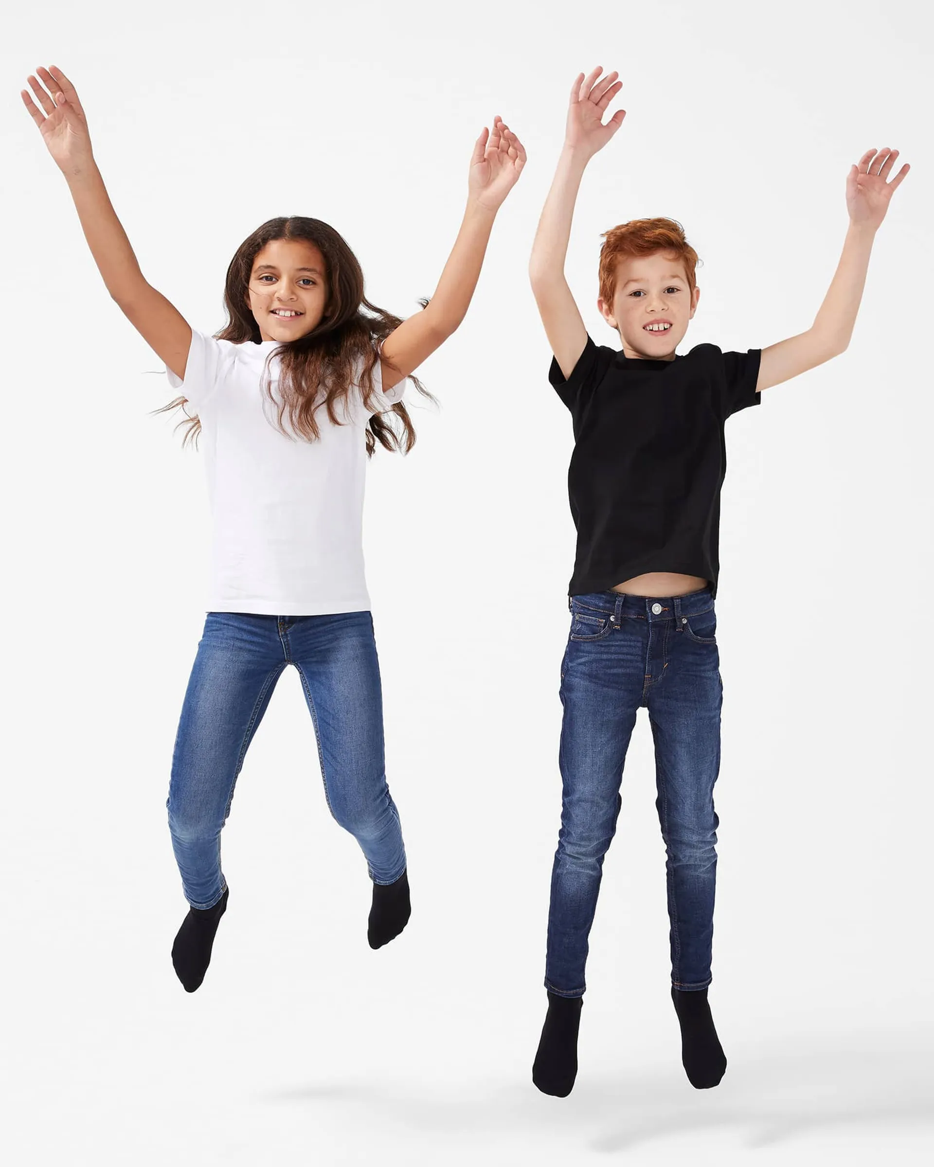 Två barn i enkla, enfärgade t-shirts, en vit och en svart, tillsammans med mörkblå jeans och svarta strumpor, hoppar upp med armarna i luften mot en ljus bakgrund.