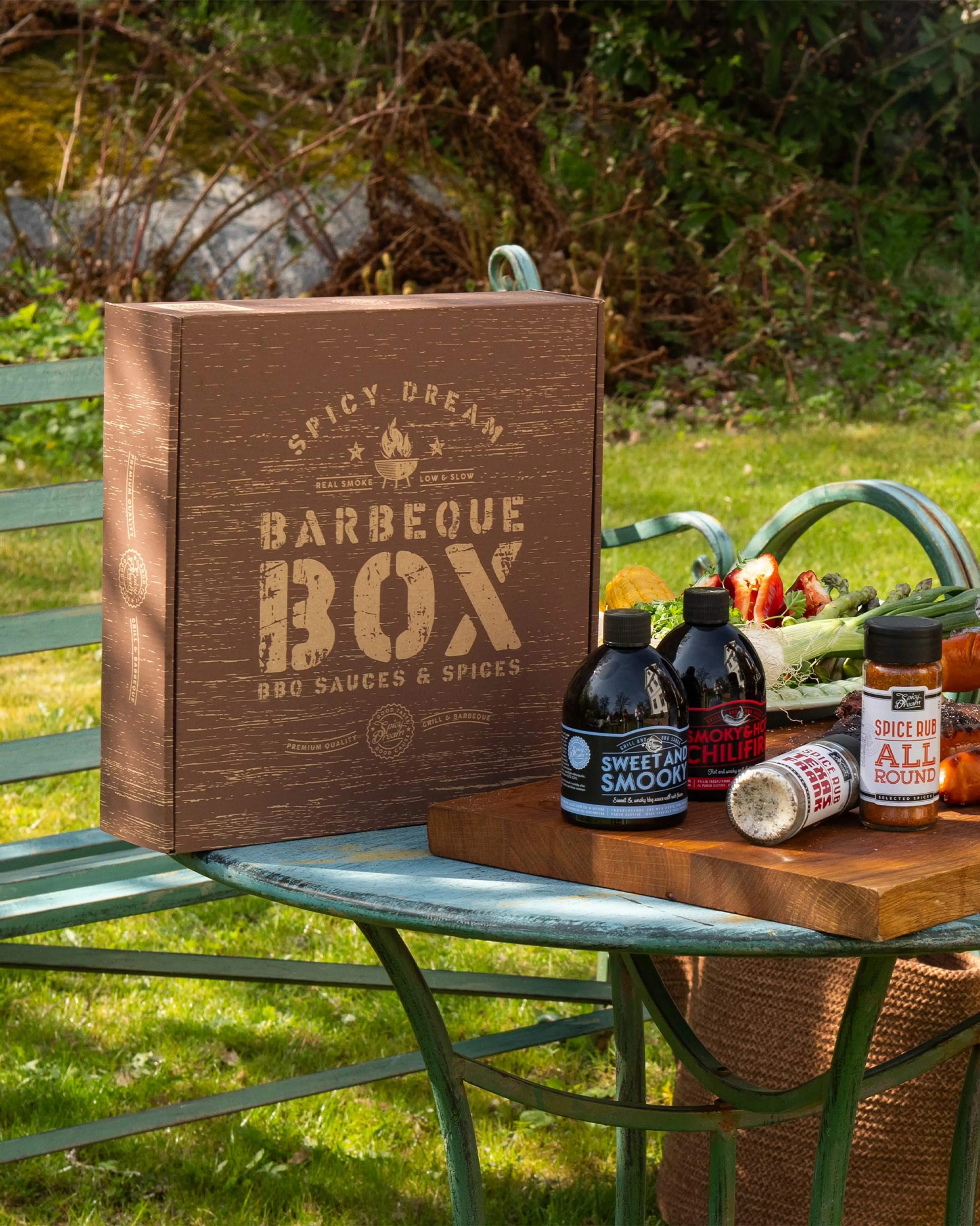 En brun box med texten "Barbeque Box" tillsammans med fyra flaskor och burkar med grillsåser och kryddor, placerade på en blågrön metallbänk utomhus. I bakgrunden syns grönska och färska grönsaker på