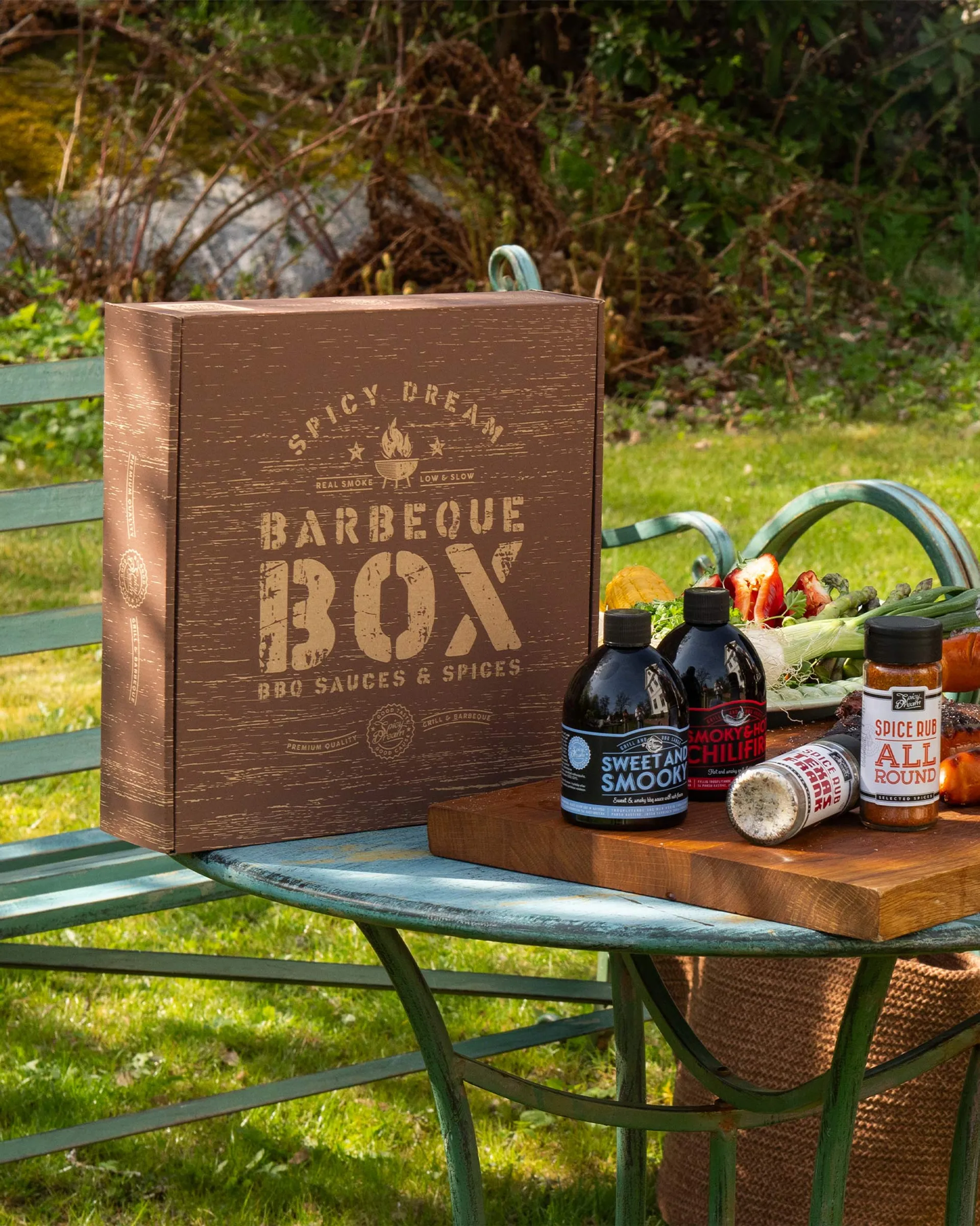En brun box med texten "Barbeque Box" tillsammans med fyra flaskor och burkar med grillsåser och kryddor, placerade på en blågrön metallbänk utomhus. I bakgrunden syns grönska och färska grönsaker på