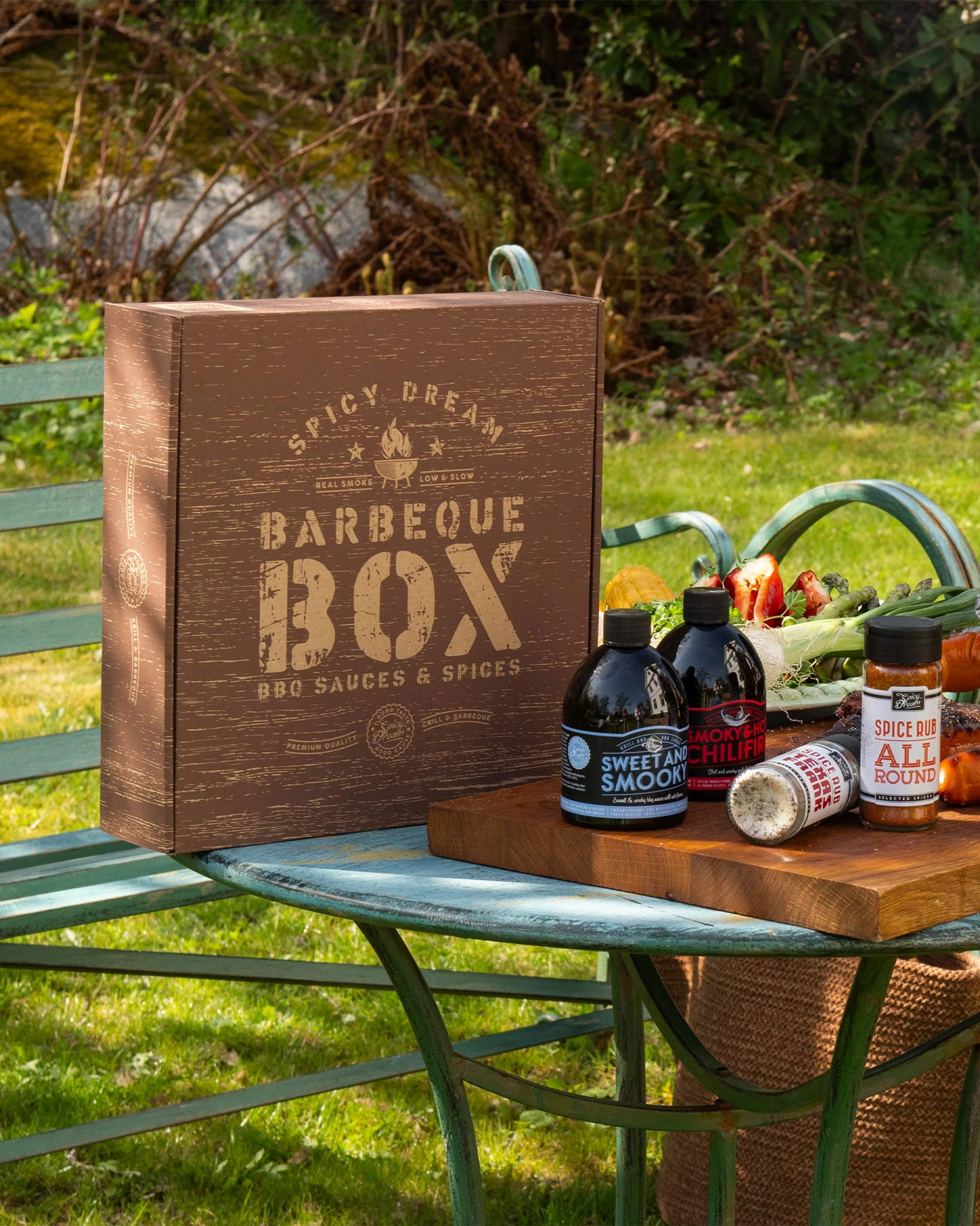 En brun box med texten "Barbeque Box" tillsammans med fyra flaskor och burkar med grillsåser och kryddor, placerade på en blågrön metallbänk utomhus. I bakgrunden syns grönska och färska grönsaker på