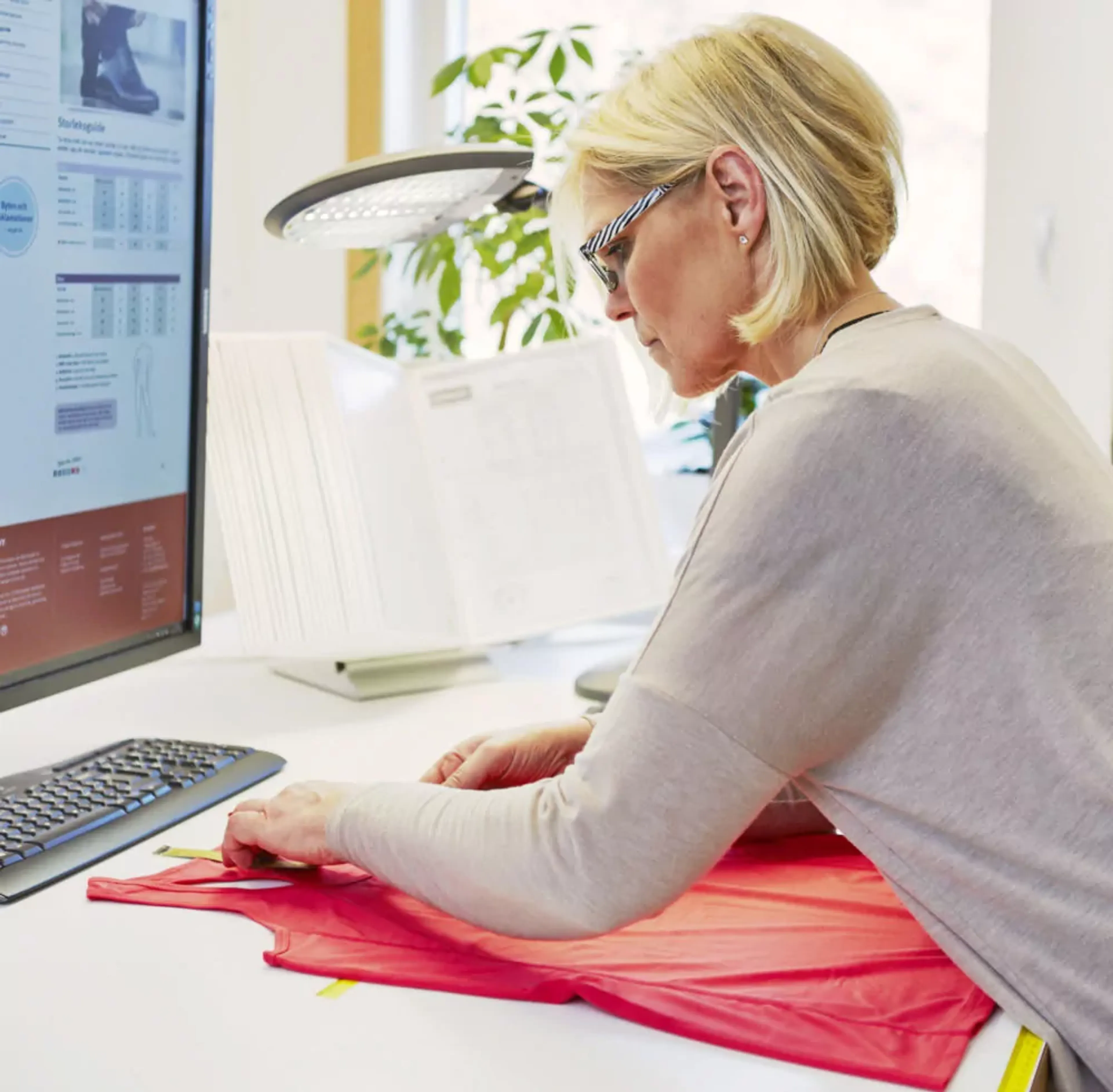 En kvinna med glasögon mäter en röd t-shirt på ett skrivbord, med en datorskärm och dokument i bakgrunden.