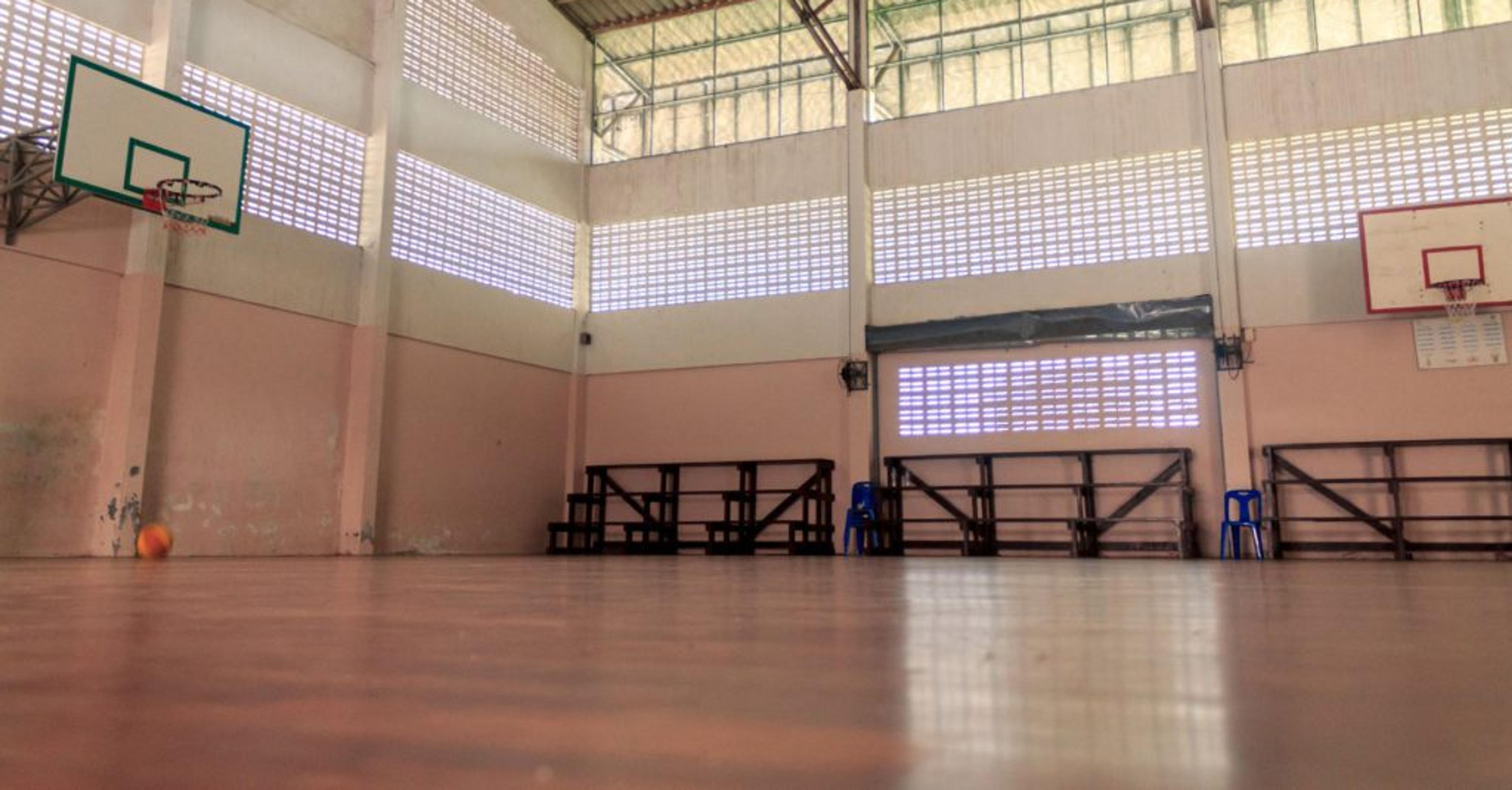 En tom gympasal med högt till tak, två basketkorgar och bänkar längs väggarna. Ljus strömmar in genom höga fönster i väggarna.