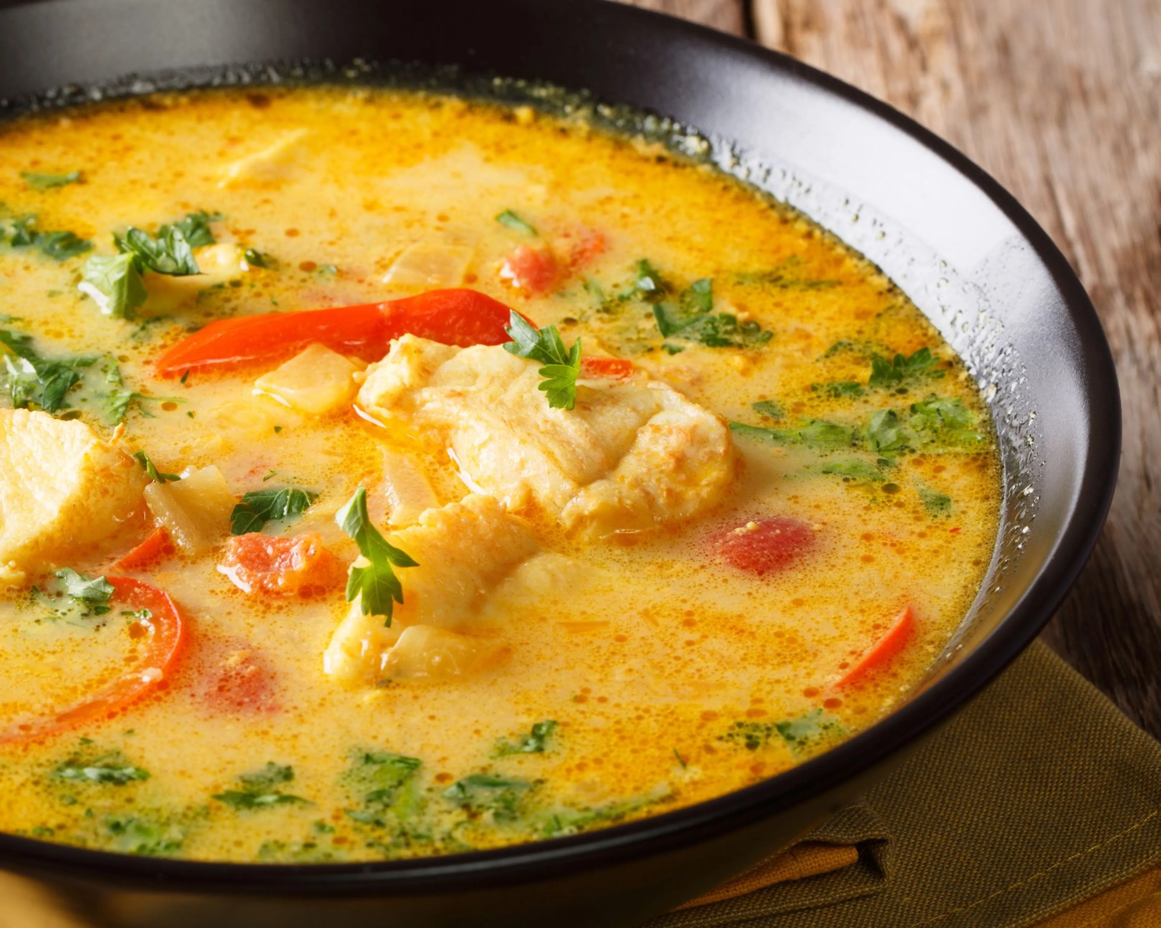 Krämig fisksoppa med saffran, röda paprikabitar och färsk persilja i en svart skål. Sopppan har en varm, gyllene färg med bitar av vit fisk.