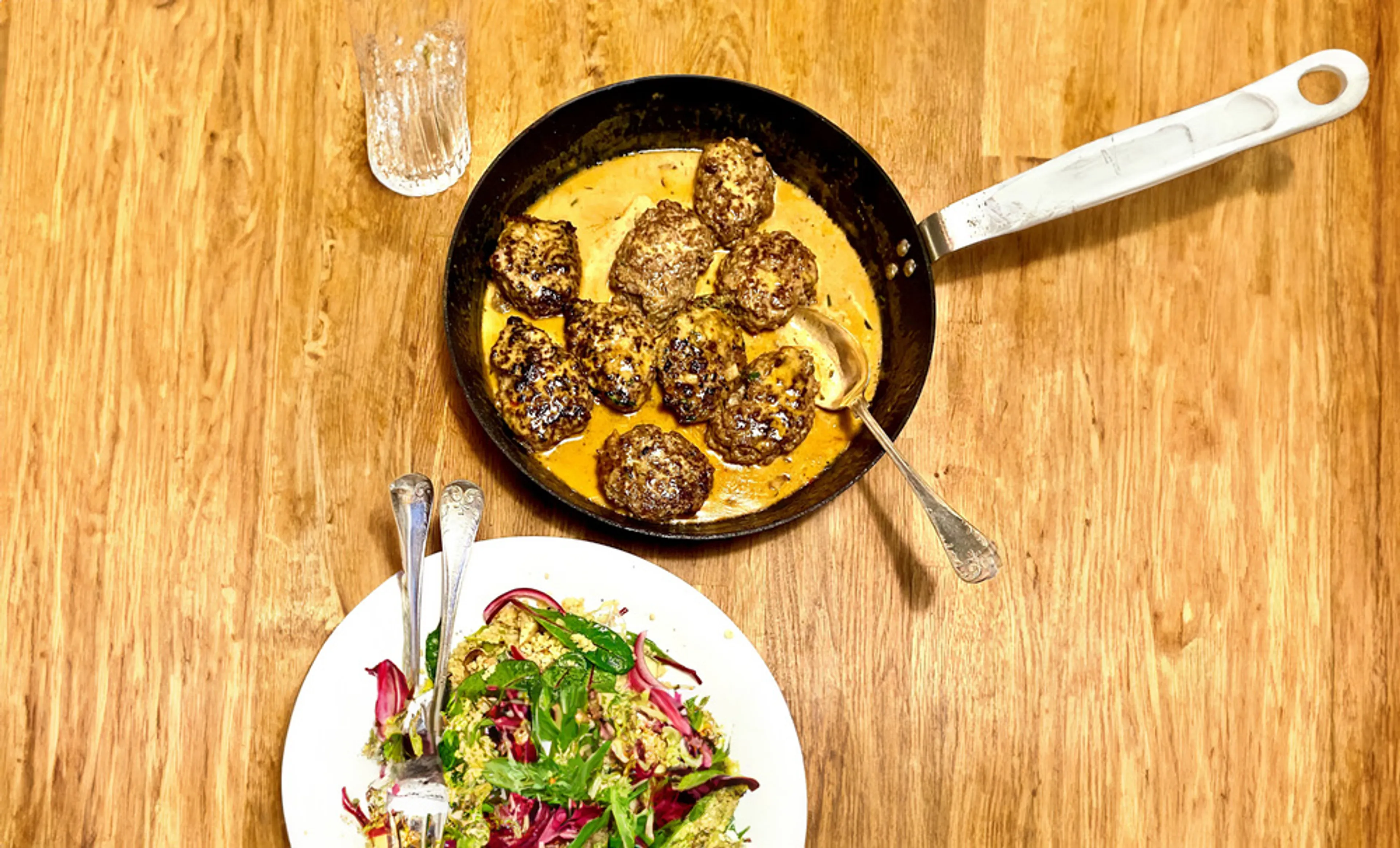 Färsbiffar som steks i en panna med krämig sås, serverade på ett träbord. En tallrik med sallad syns i förgrunden.