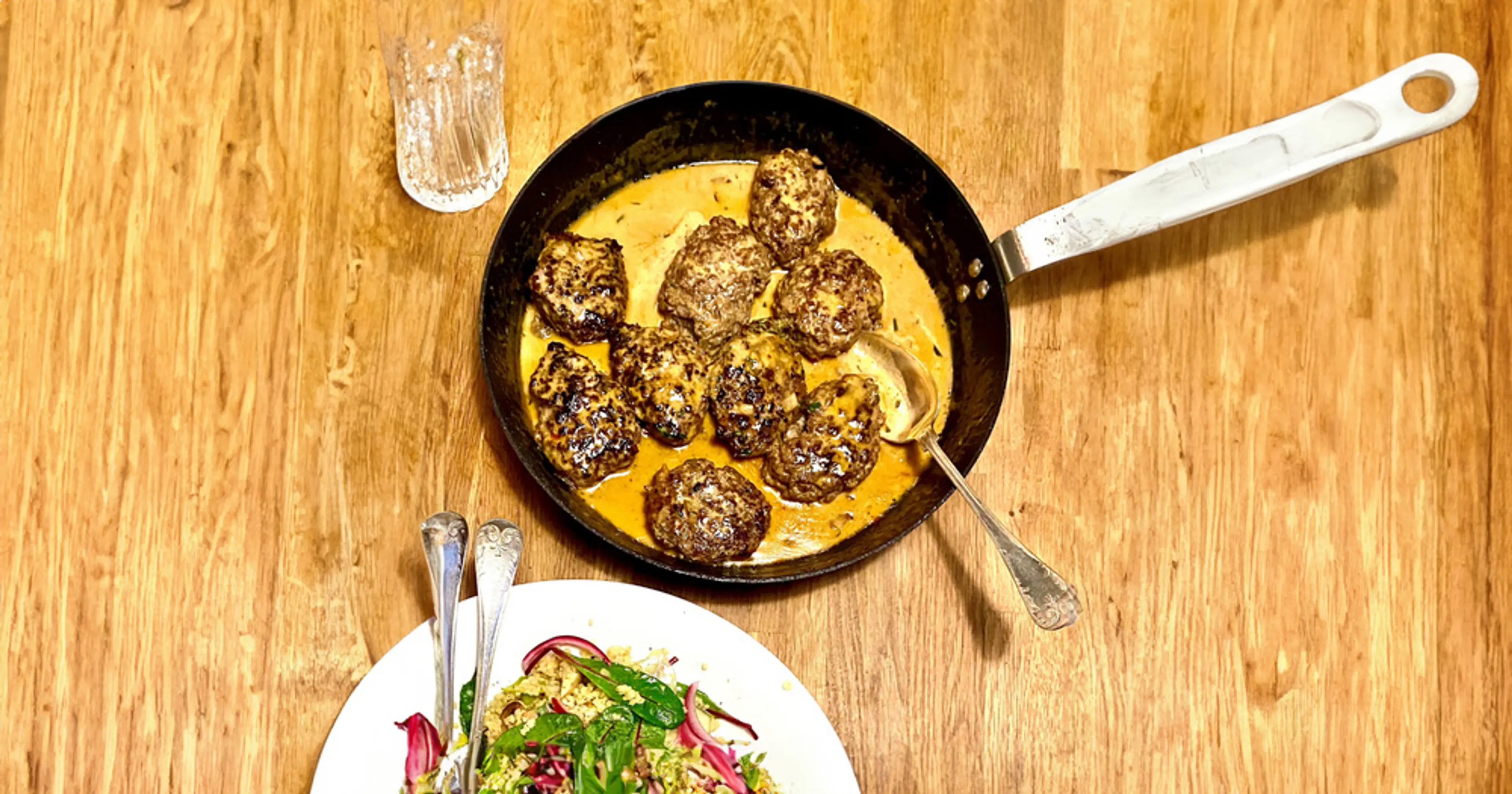 Färsbiffar som steks i en panna med krämig sås, serverade på ett träbord. En tallrik med sallad syns i förgrunden.