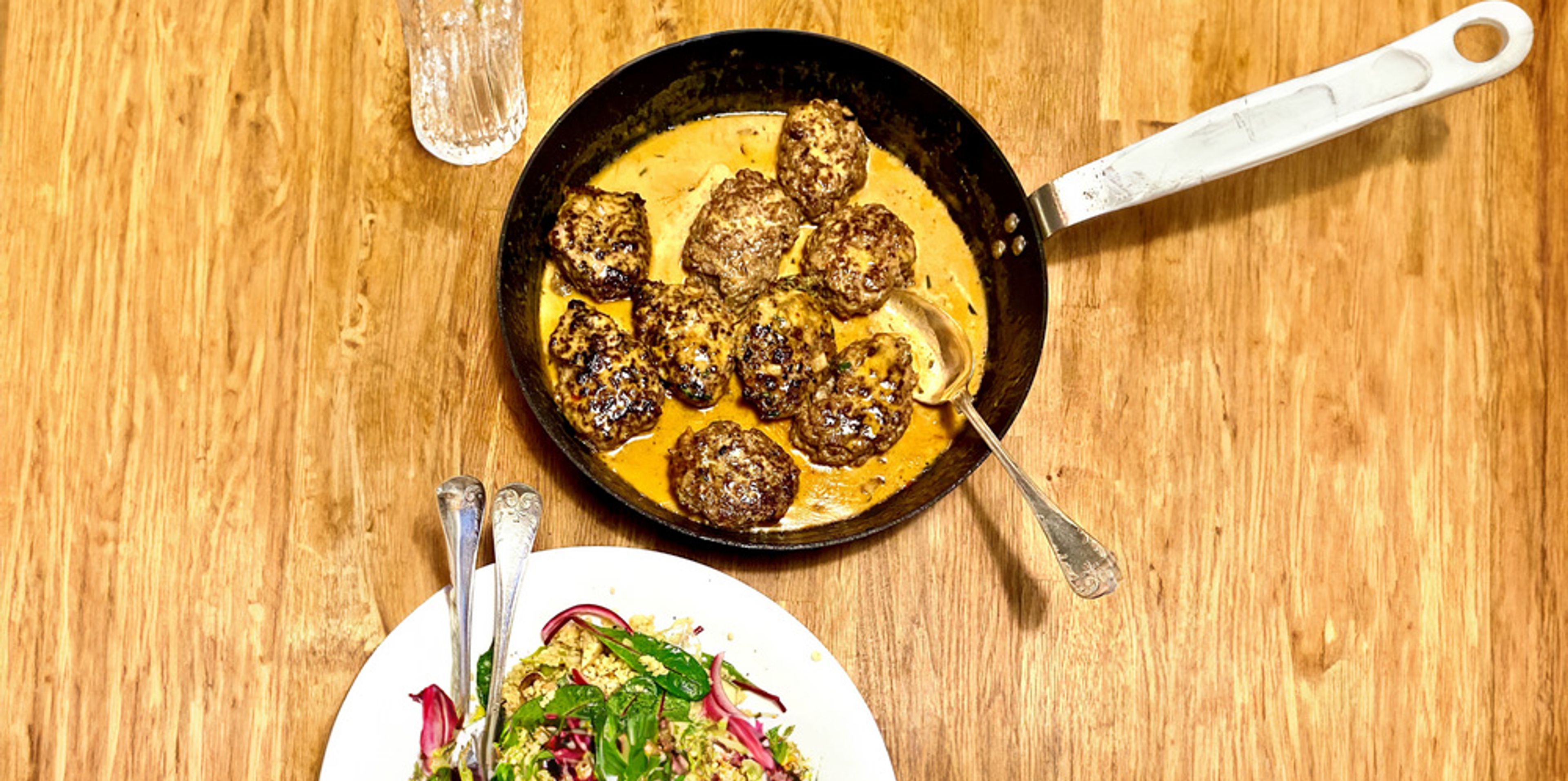 Färsbiffar som steks i en panna med krämig sås, serverade på ett träbord. En tallrik med sallad syns i förgrunden.