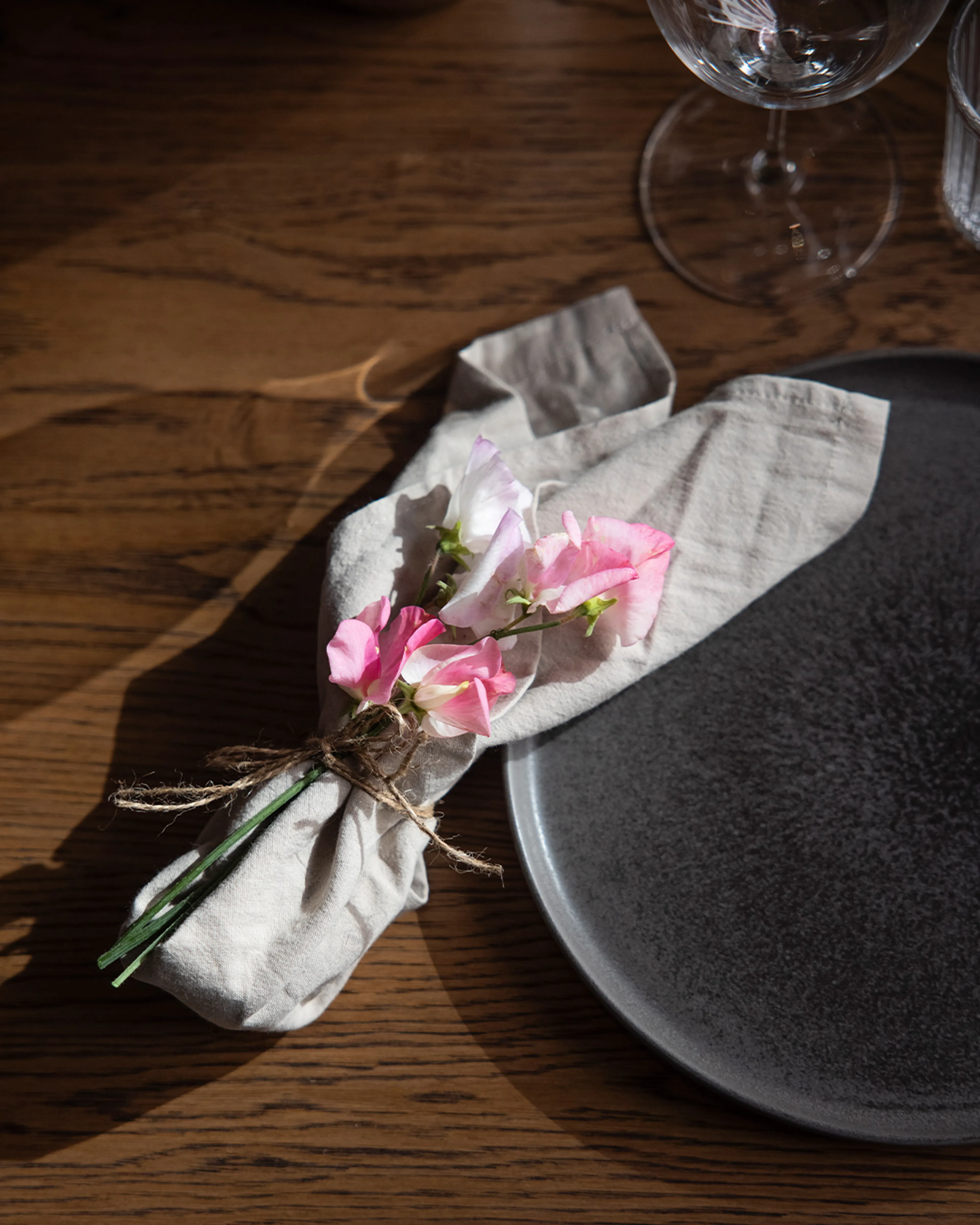 En linneservett knuten med snöre och dekorerad med små rosa blommor, placerad bredvid en mörk tallrik på ett träbord. En elegant och avskalad dukning i naturliga färger.