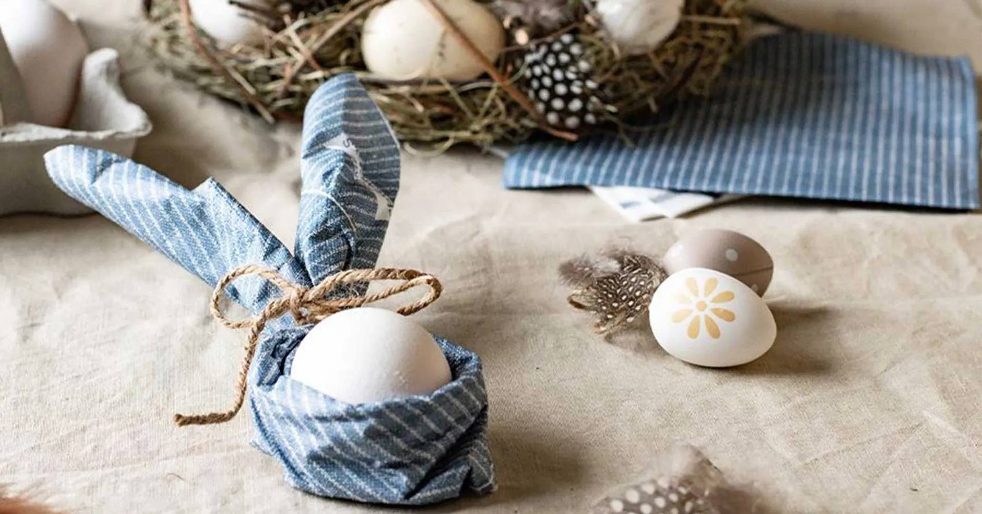 Ett vitt ägg insvept i en blå och vitrandig servett knuten som kaninöron, omgiven av fjädrar och ett dekorativt fågelbo med ägg. Bakgrunden består av en ljus duk och fler blå servetter.