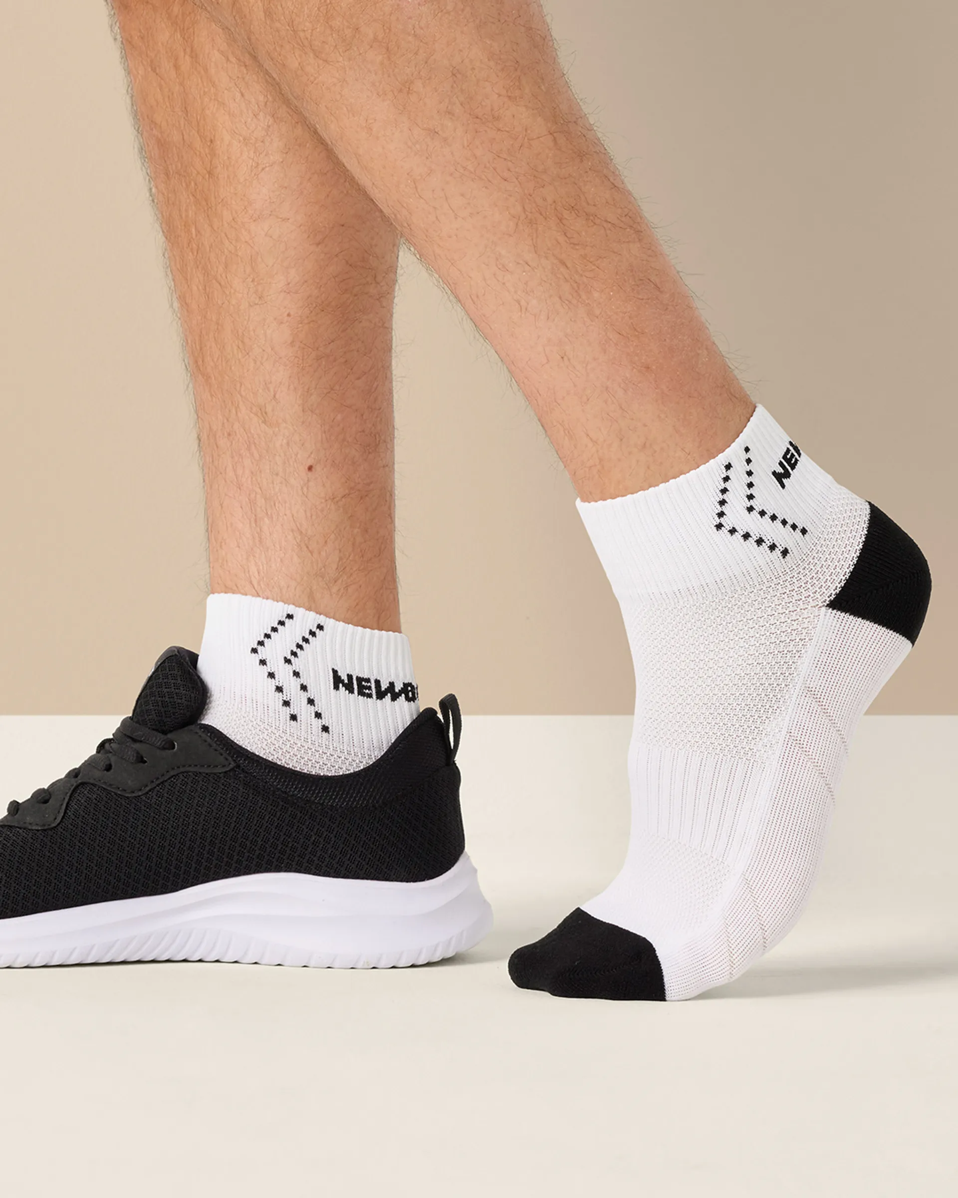 Ett par vita strumpor med svart tå- och hälparti, designade för prestation och komfort, visas på en mans fot bredvid en svart sportig sko. Strumporna har också svart mönster och text vid skaftet.